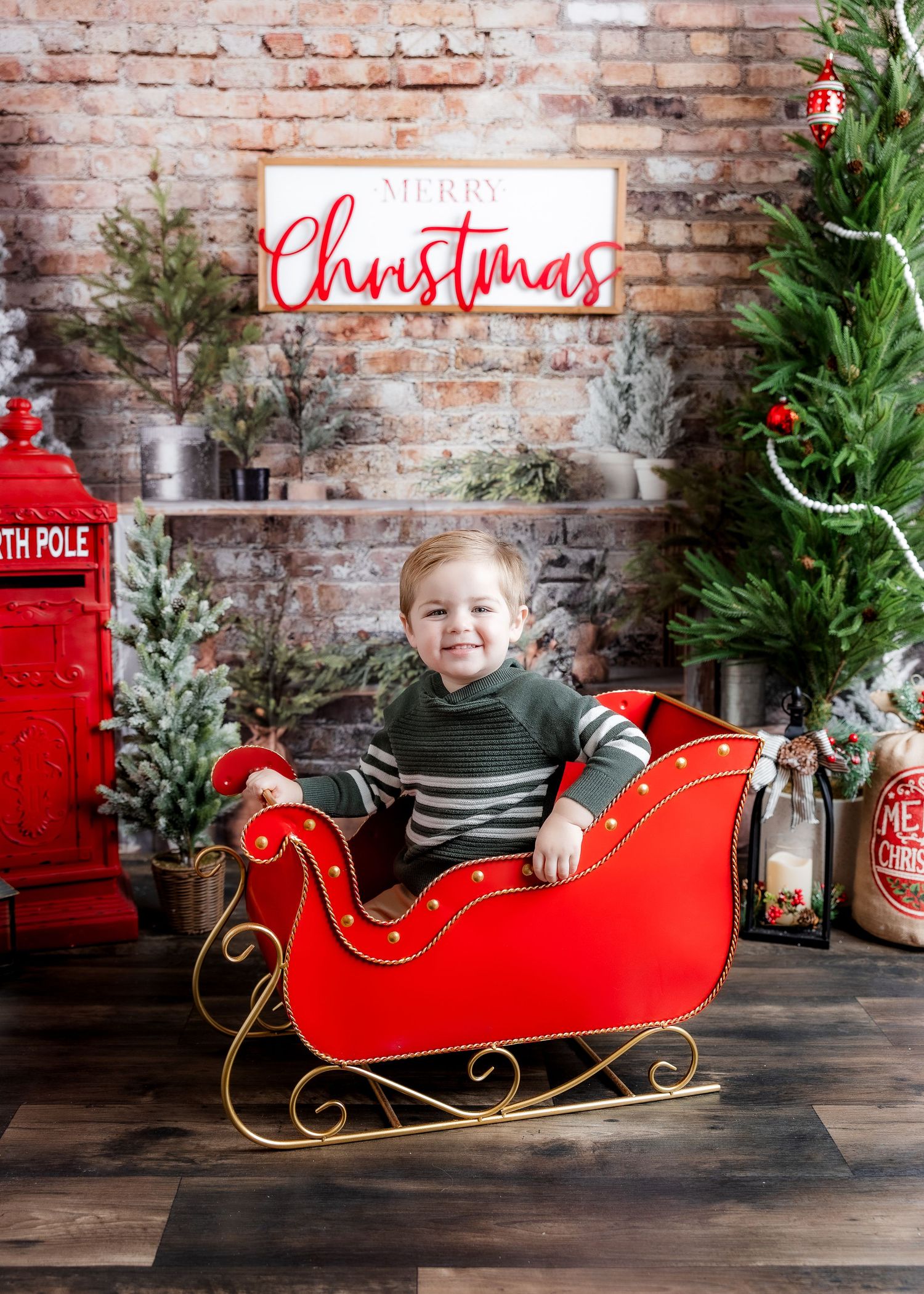Cheerful toddler boy in striped sweater sitting in red Christmas sleigh with Merry Christmas sign.
