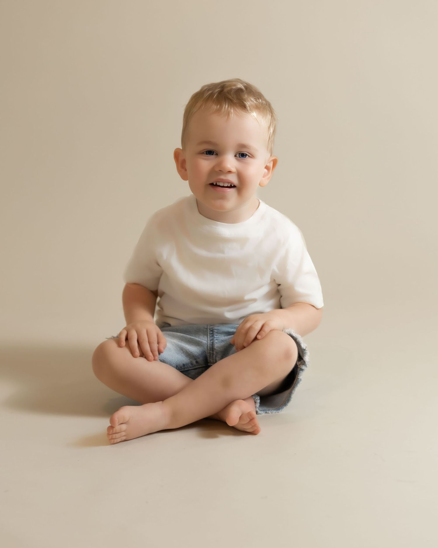 Blonde toddler boy in white tee and denim shorts sits cross-legged smiling on a neutral background.