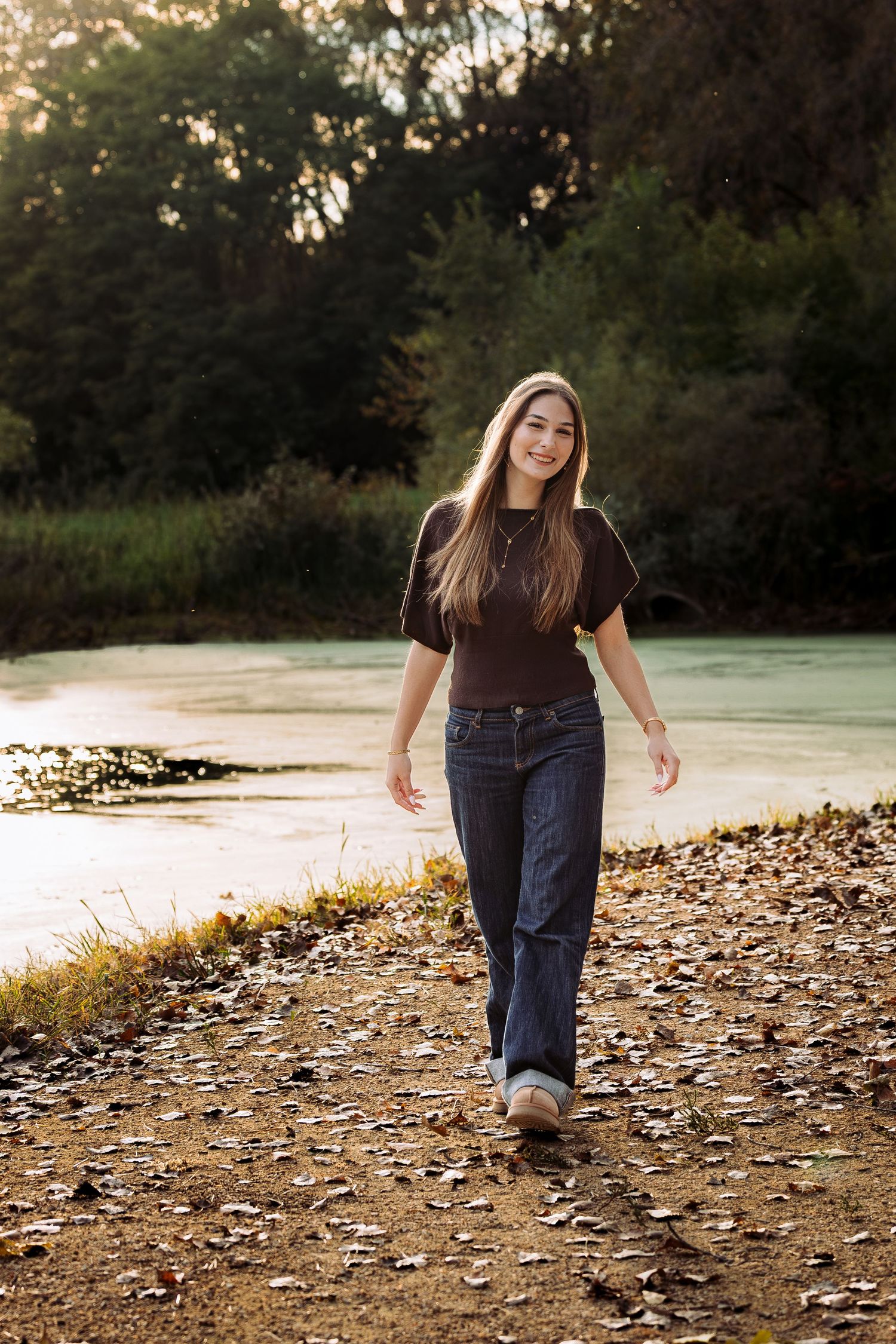 Young woman in dark jeans and brown top walking along a leaf-covered path beside a serene pond in autumn.