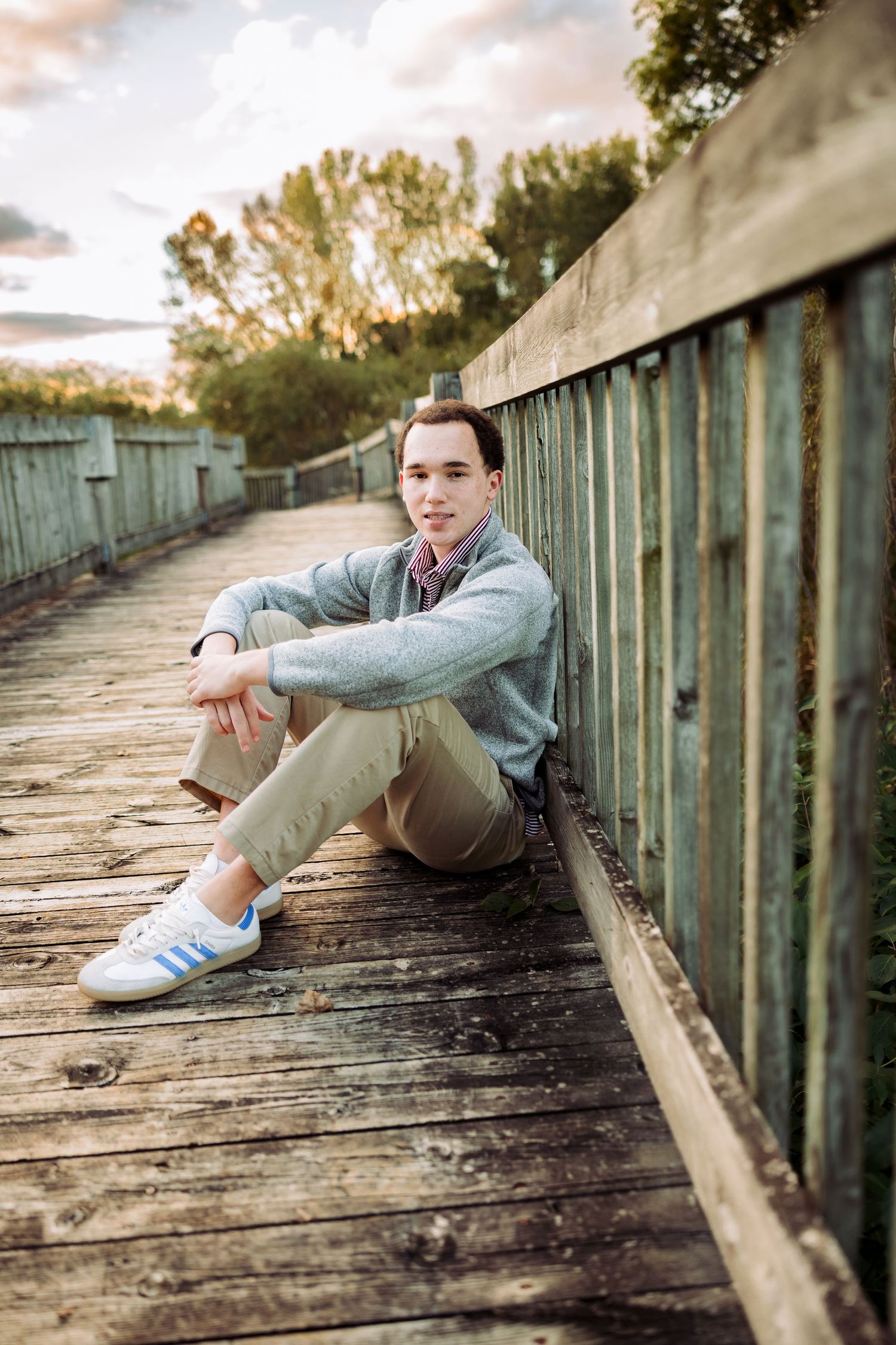 Young man in gray sweater and khakis sits on wooden bridge deck, leaning against fence at golden hour.