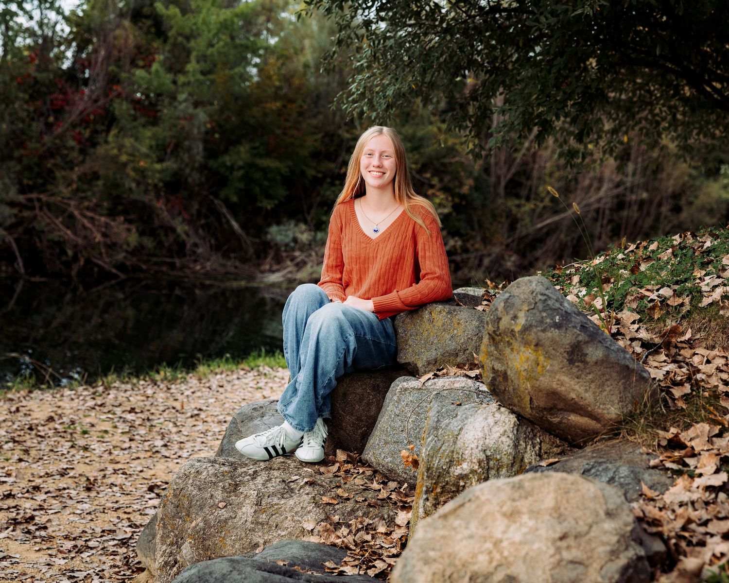 A smiling teenage girl with blonde hair sits on rocks wearing an orange sweater and jeans in an outdoor fall setting.
