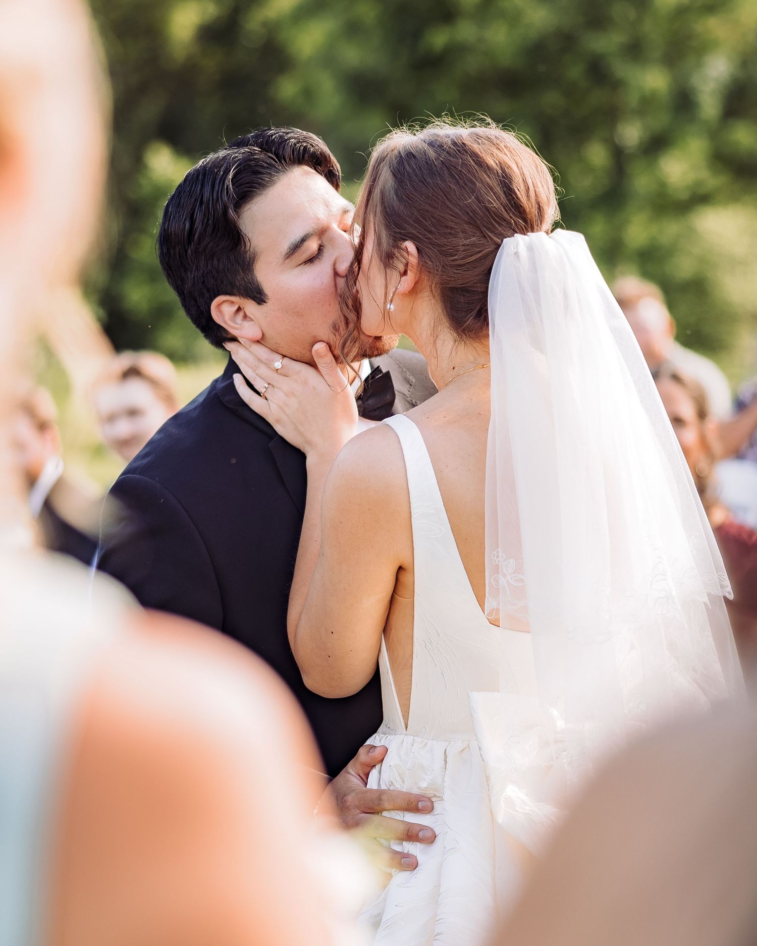 Bride and groom share their first kiss at an outdoor wedding ceremony, bride wearing a white gown and veil.