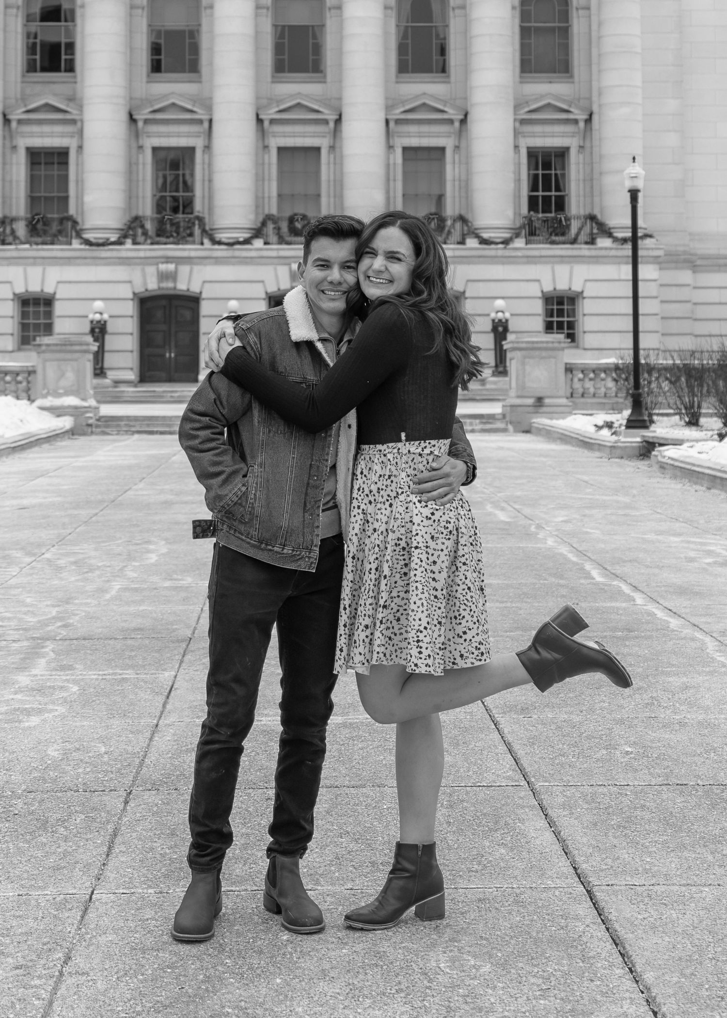 Black and white photo of a couple embracing joyfully outside a grand capitol building in winter.