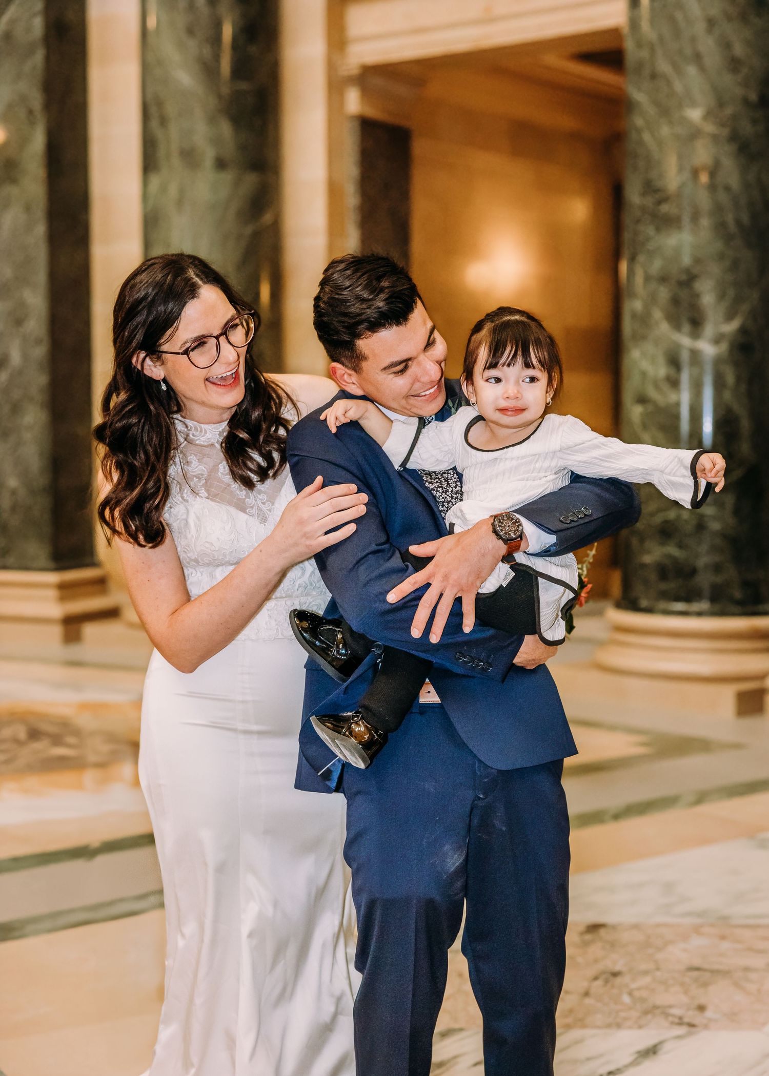 Couple in wedding attire holding a joyful toddler with arms outstretched in a grand marble hall.