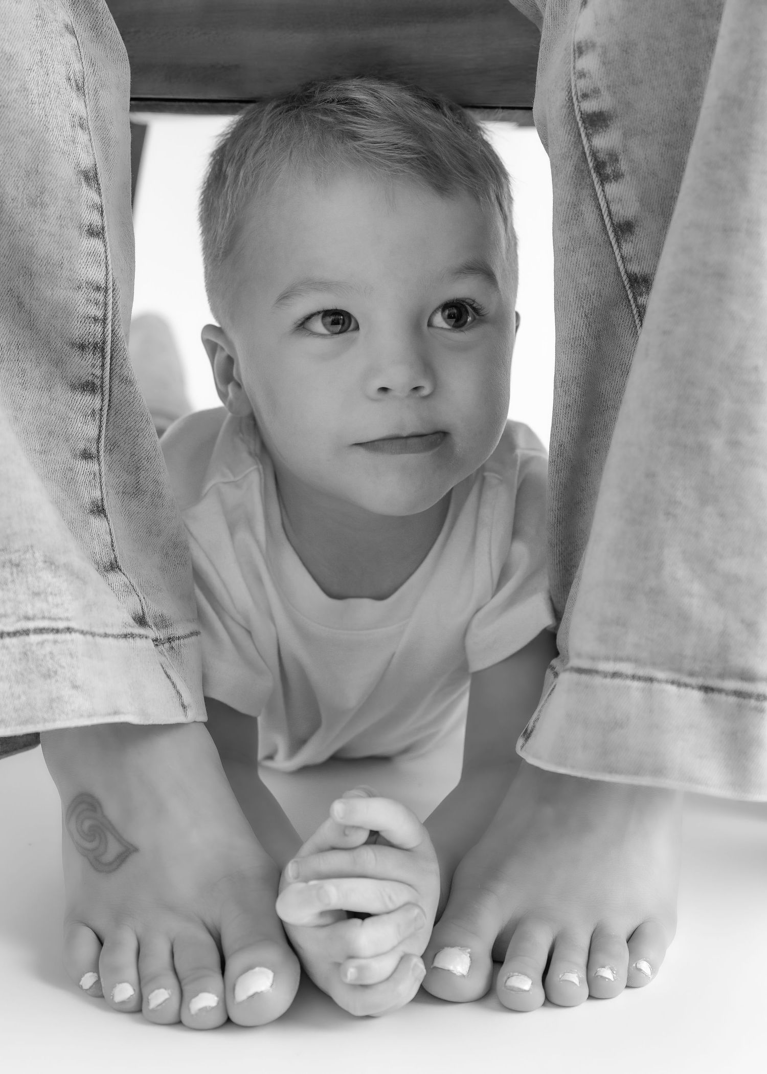 Curious baby boy peeking between parents legs in charming black and white family portrait.
