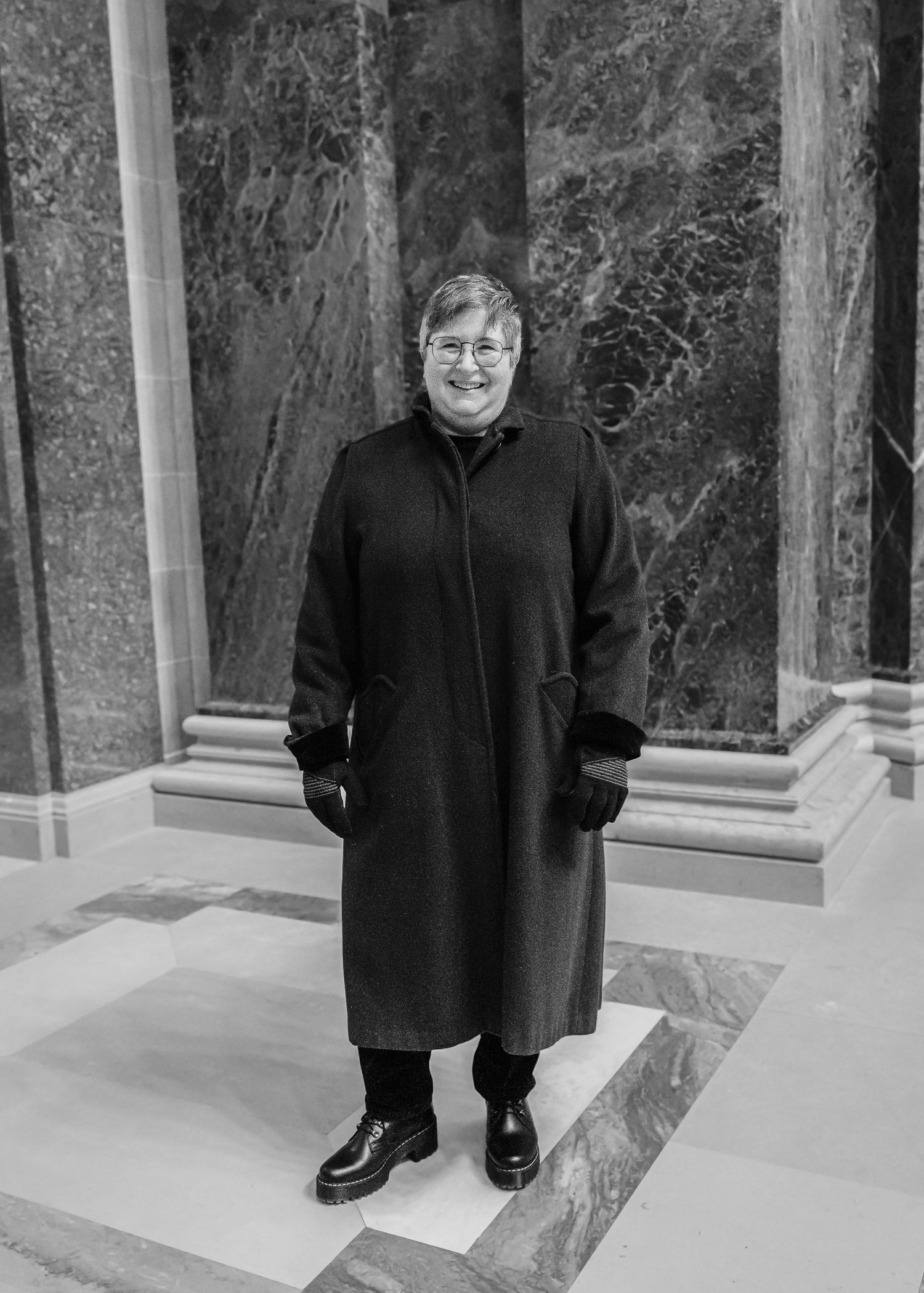 A woman in a long black coat and gloves stands between marble columns in a black and white photo.
