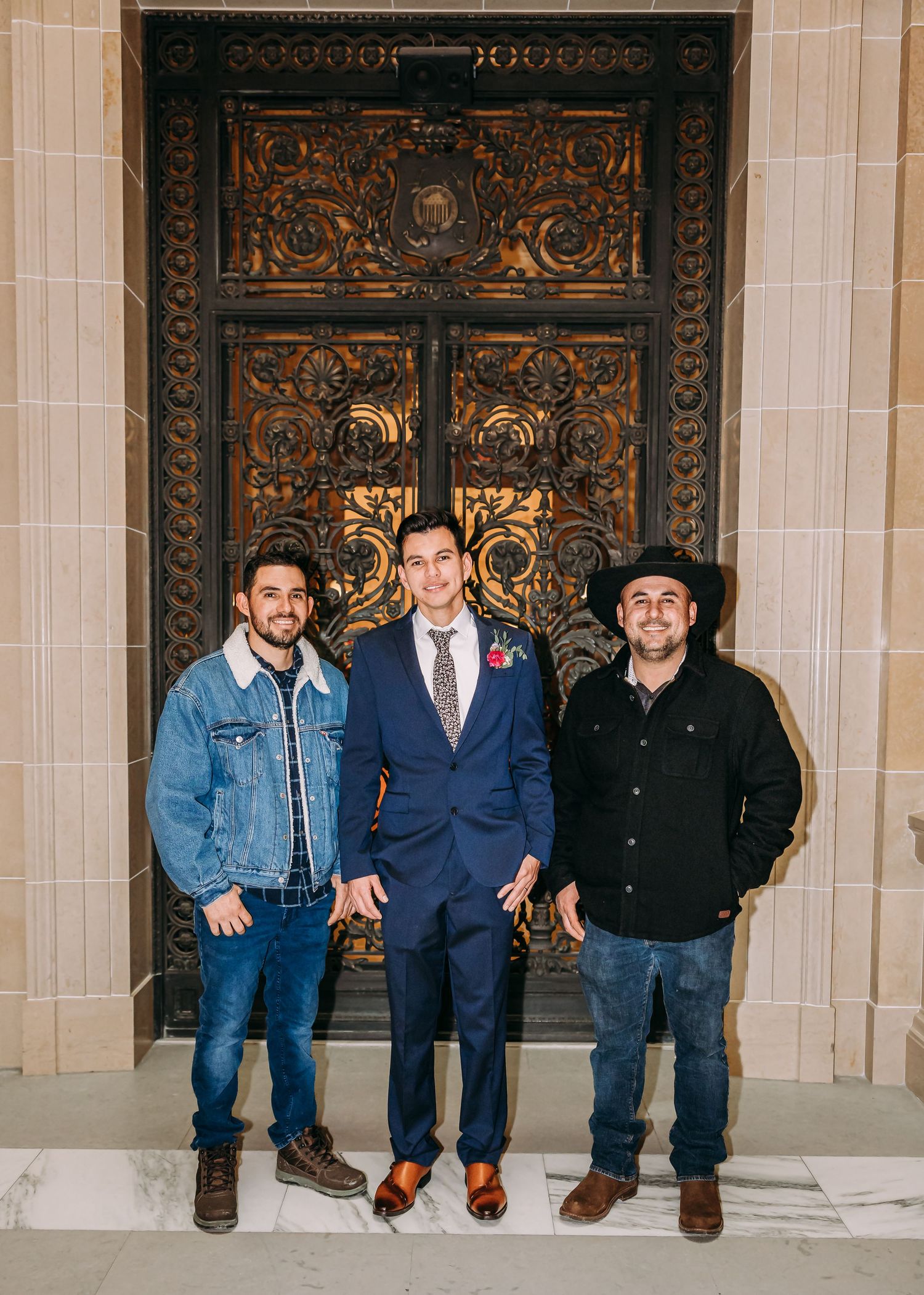 Three men posing in front of ornate bronze doors, center man wearing blue suit with floral boutonniere.