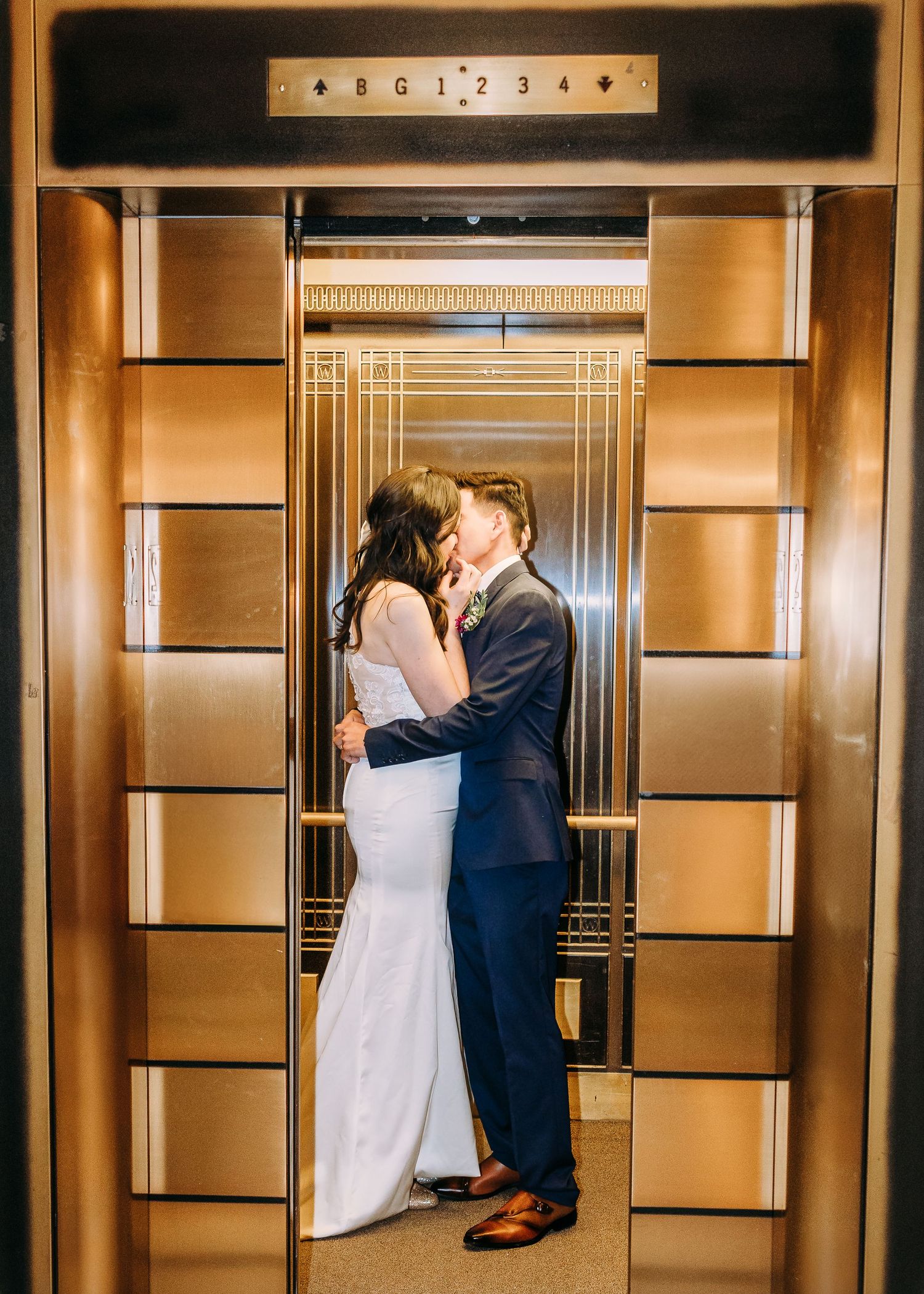 Bride and groom share a tender kiss inside an elegant gold Art Deco elevator with decorative panels.