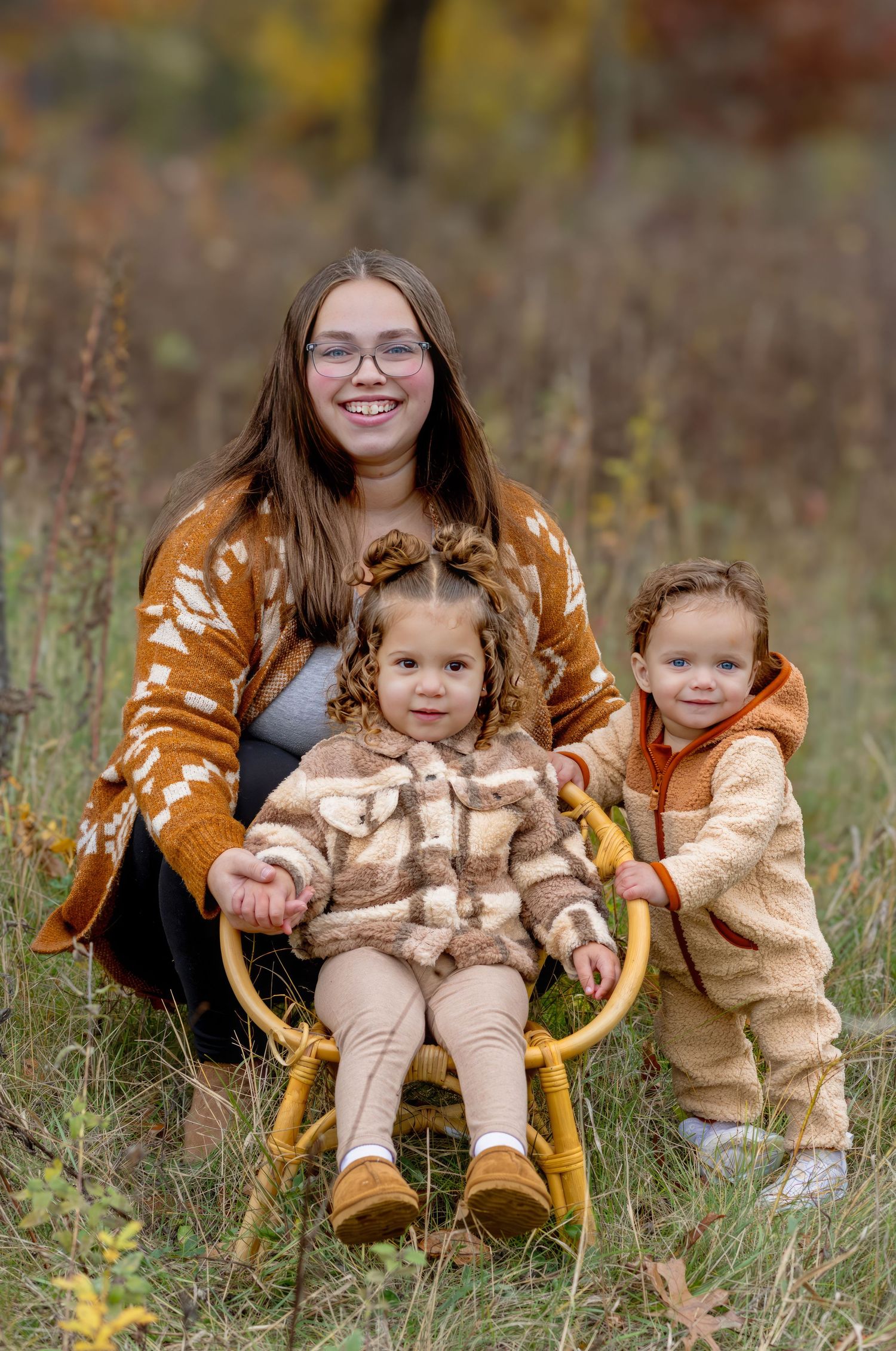A smiling woman in an orange cardigan poses with two young children in a field during autumn.