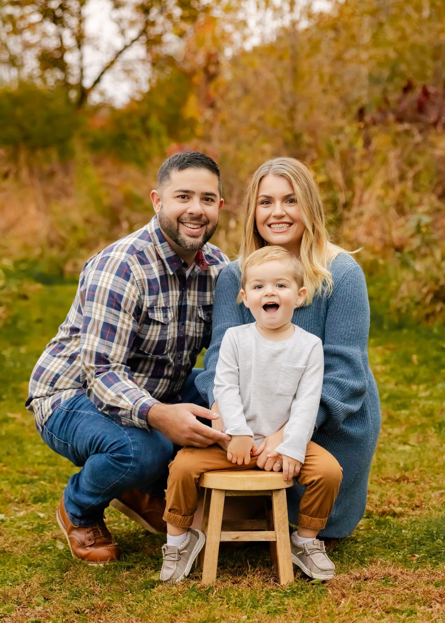 Happy family of three posing outdoors in autumn, parents smiling with excited toddler on wooden stool.