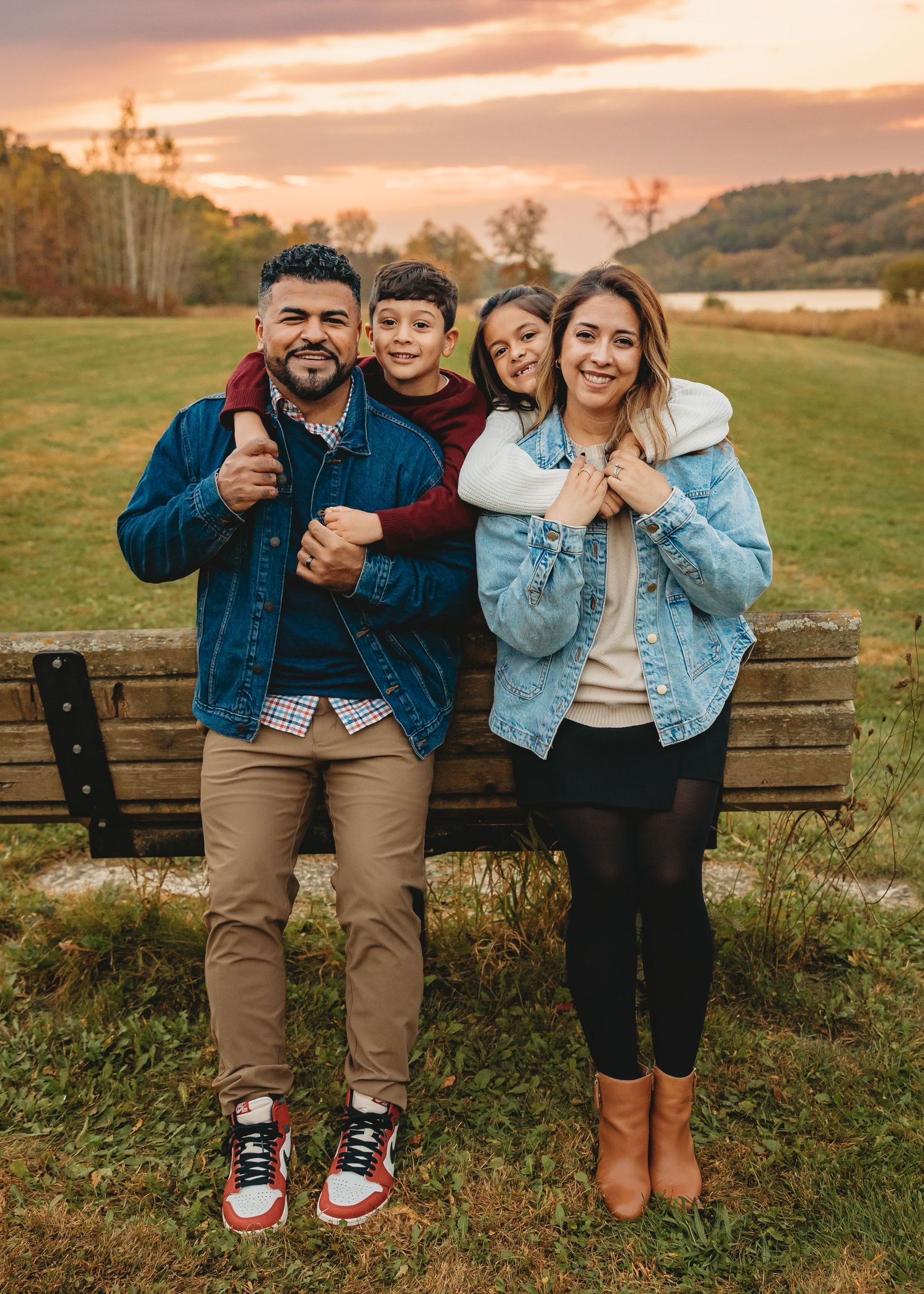 A joyful family of four poses together on a bench during a colorful autumn sunset.