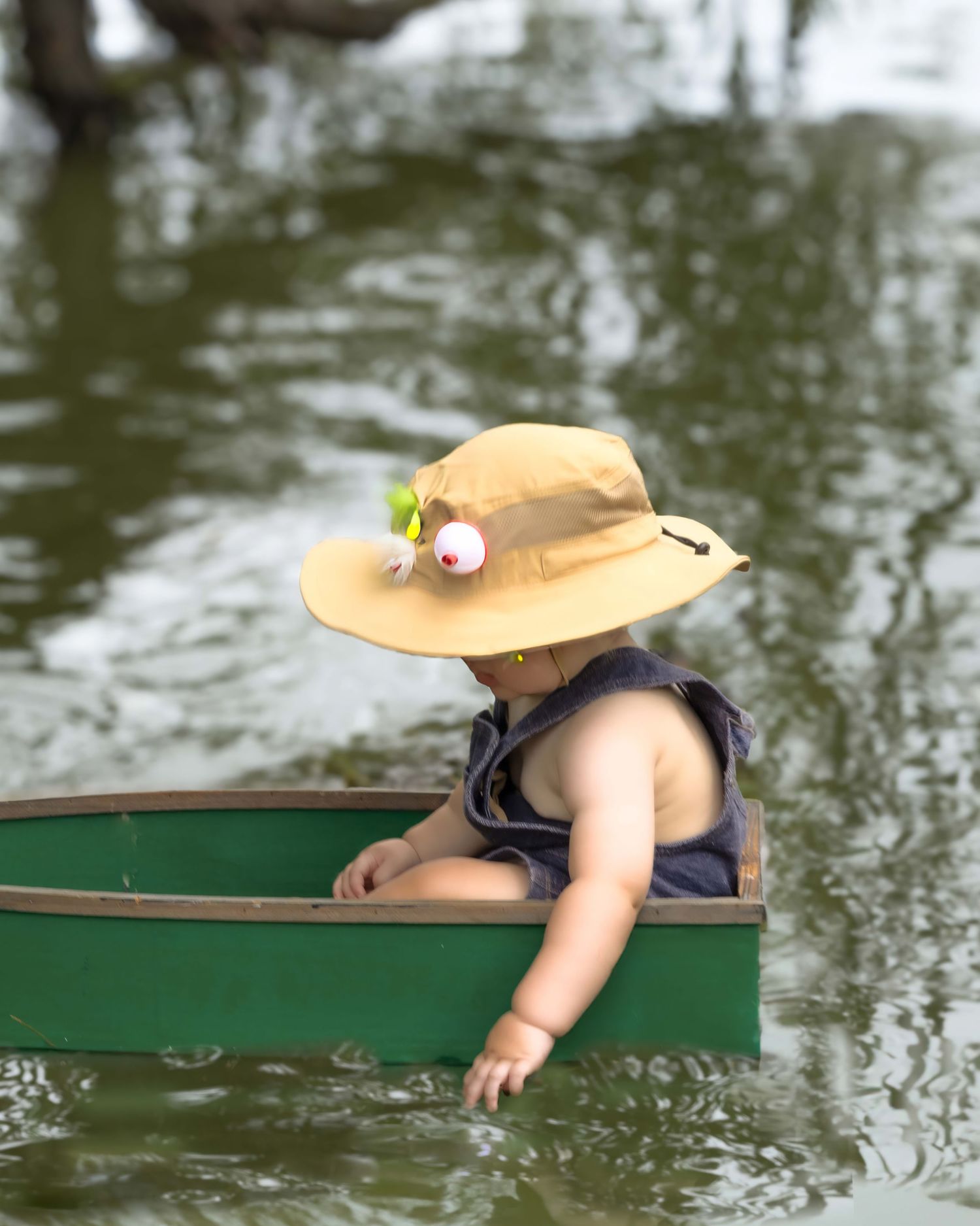 Adorable baby wearing sun hat sitting in green canoe on calm water.