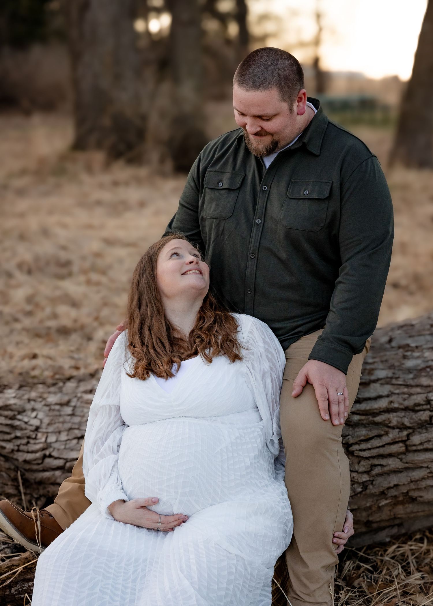 Pregnant woman in white dress sitting on log with partner in green shirt at golden hour.