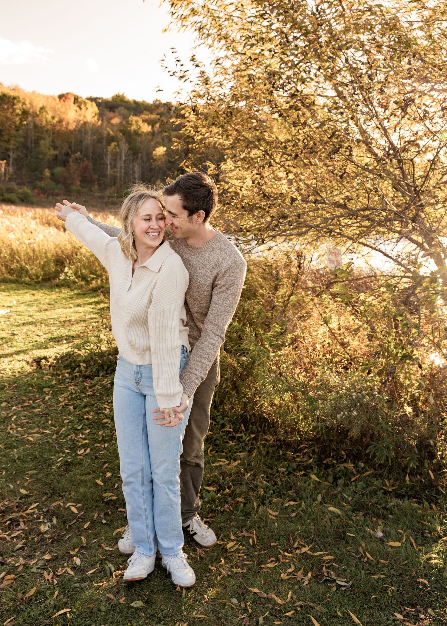 Man kissing woman on cheek while she laughs outdoors near golden fall foliage.