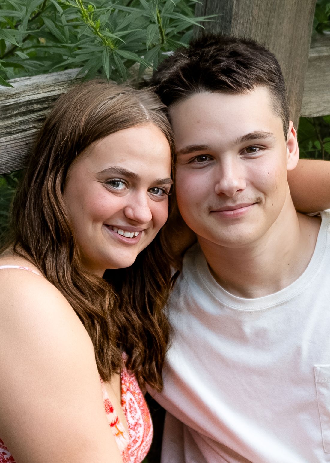 Smiling teenage couple posing closely together outdoors leaning against a wooden fence in summer.