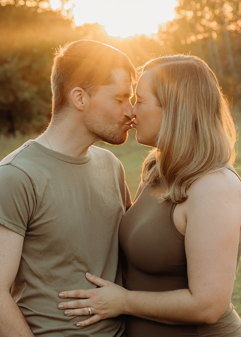 Couple sharing a tender kiss at golden hour sunset in a lush green field.