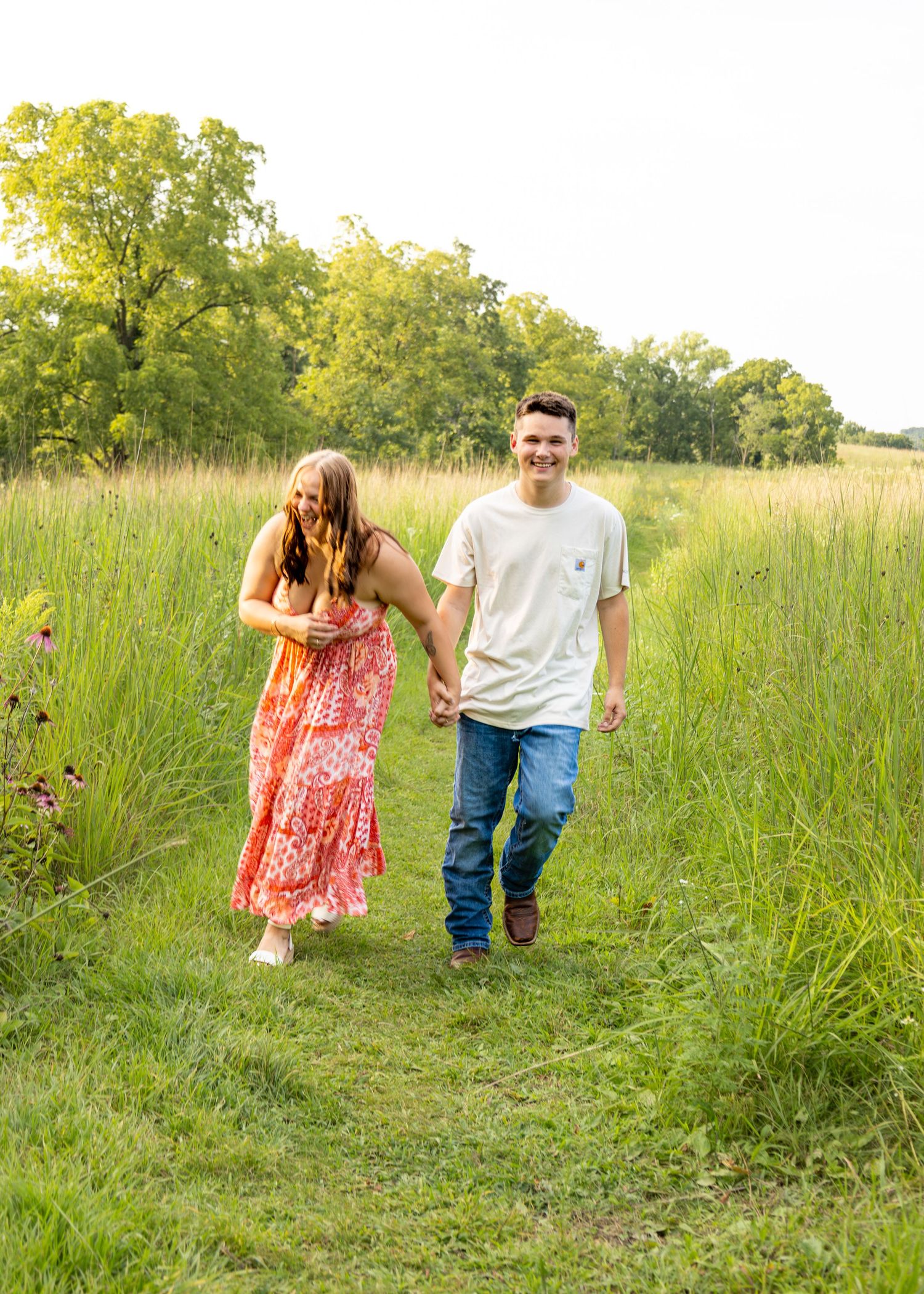 A smiling young man leads his laughing partner by the hand along a sunny green meadow path.