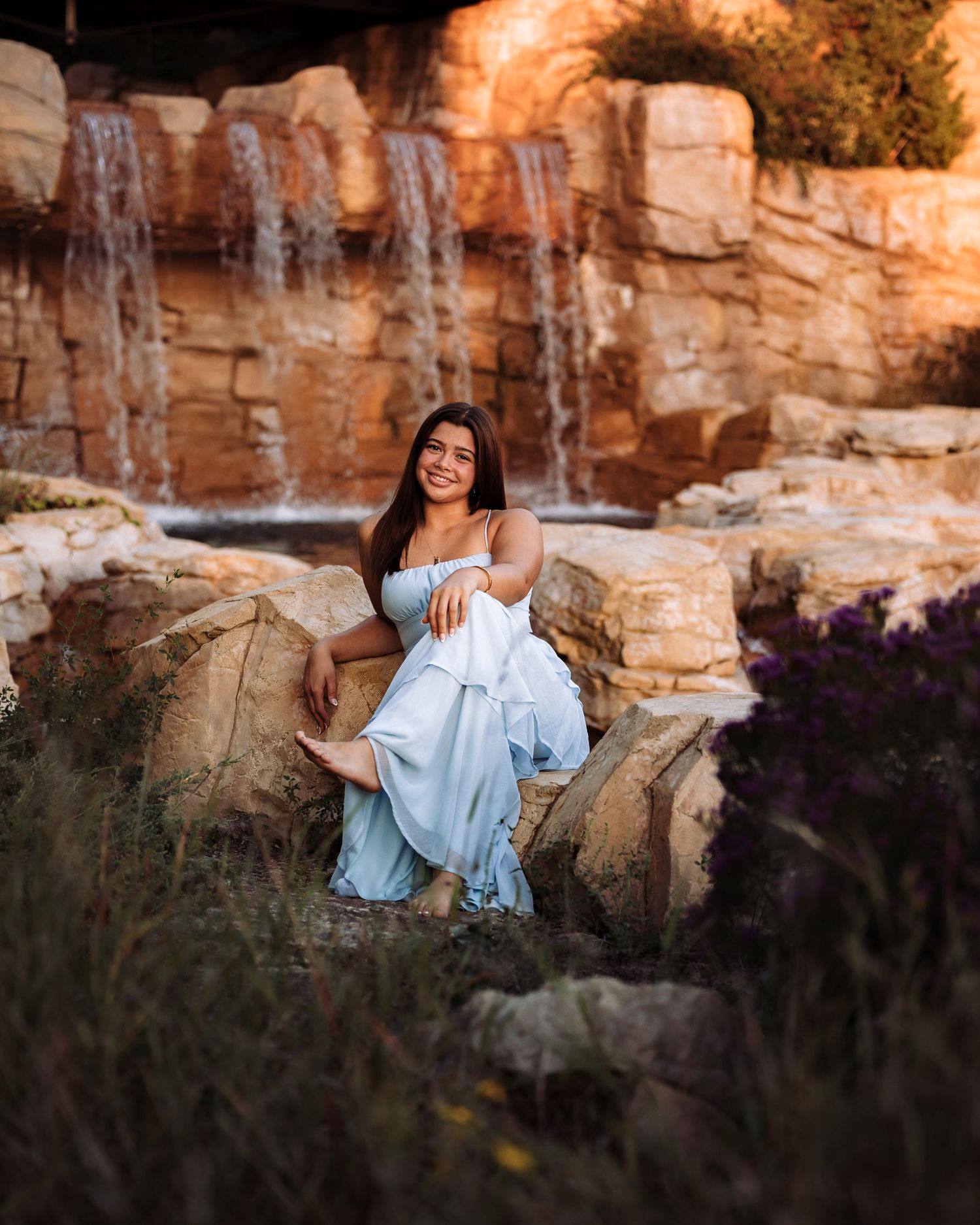 A smiling woman in a light blue dress sits on rocks near a waterfall surrounded by purple wildflowers at golden hour.
