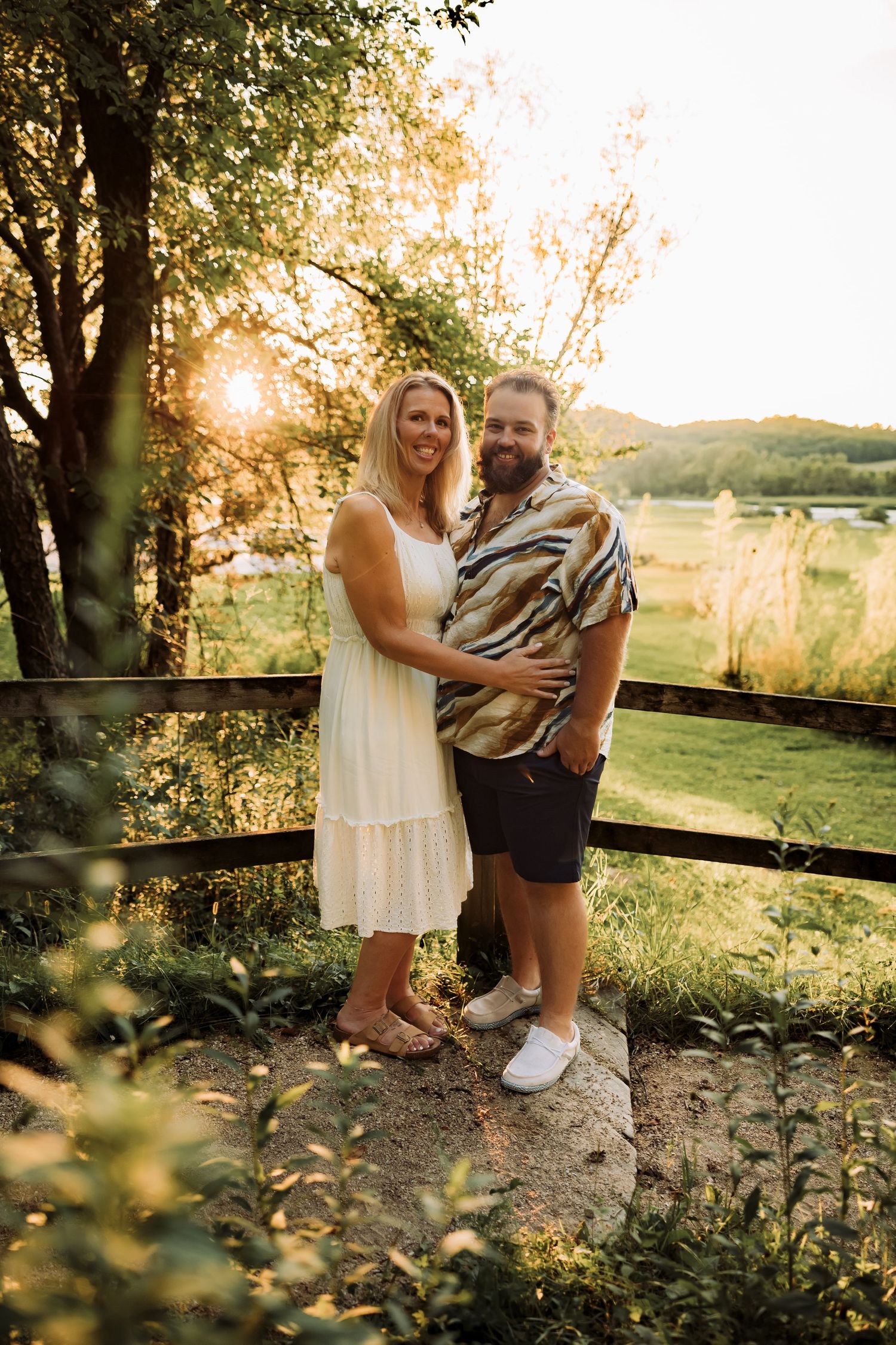 Couple embracing at golden hour sunset near wooden fence with lush green landscape and lake in background.