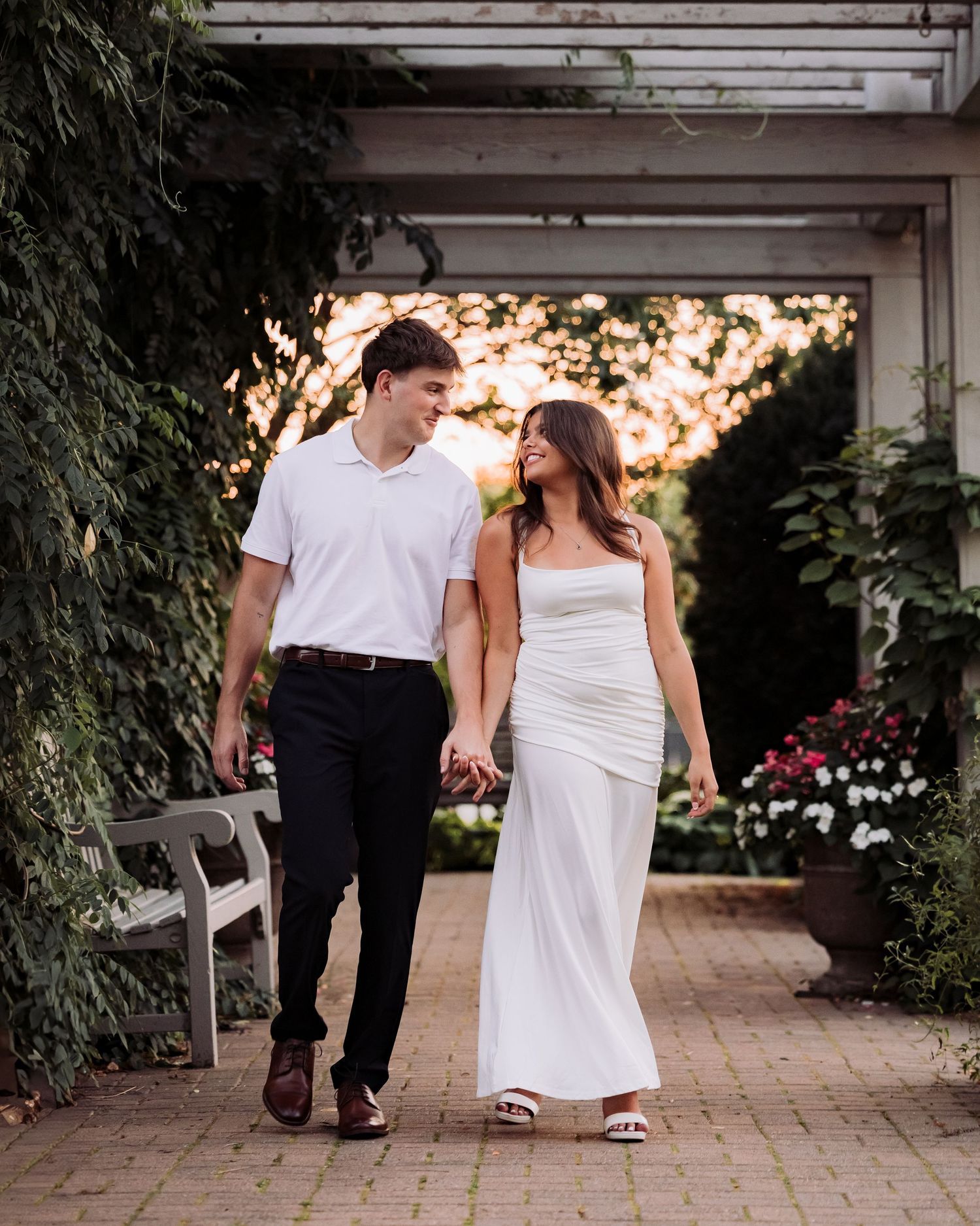 Couple in white outfits holding hands walking through a garden pergola with lush greenery and flowers.
