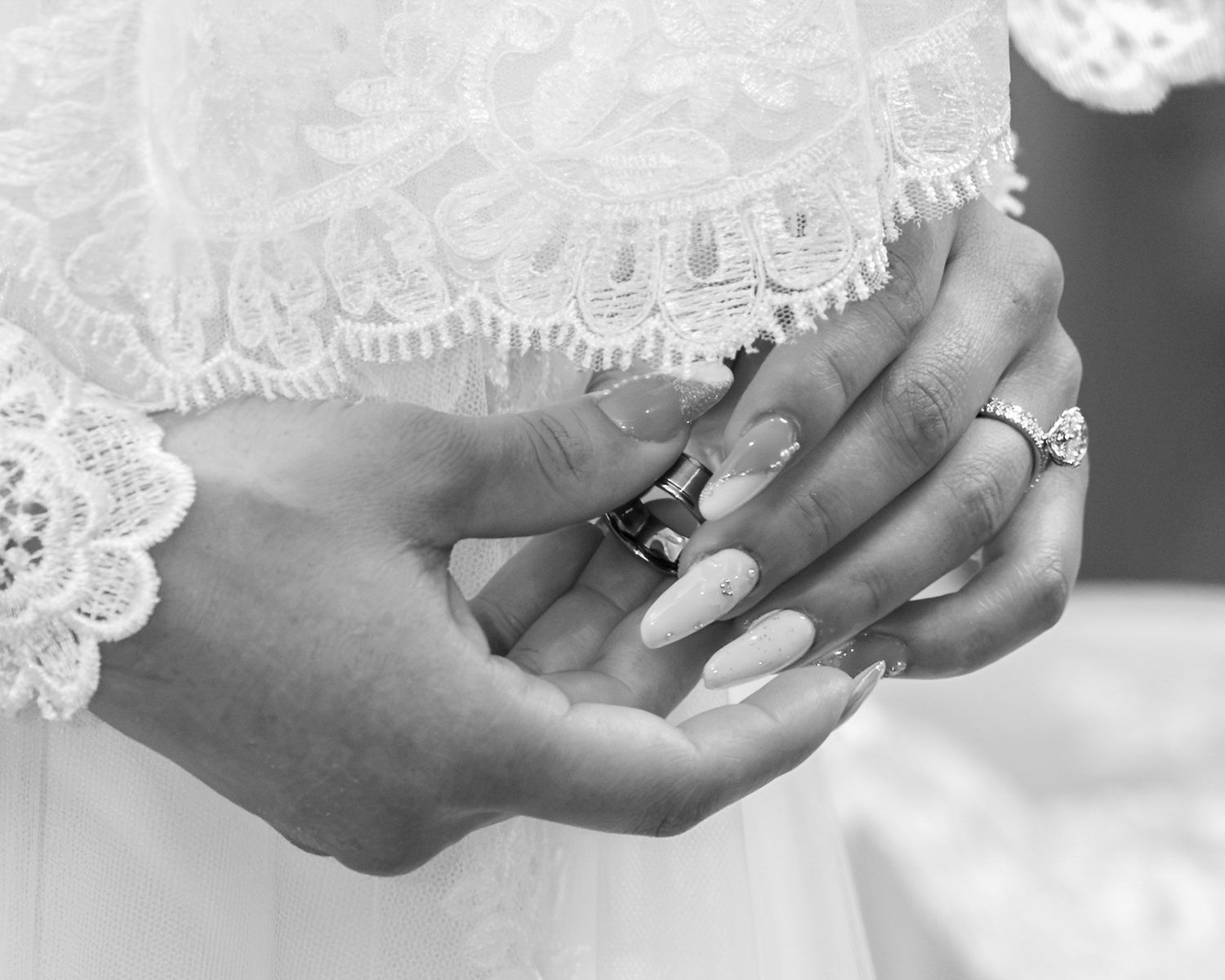Black and white close-up of a bride's hands with long manicured nails holding a wedding ring, wearing a lace dress.