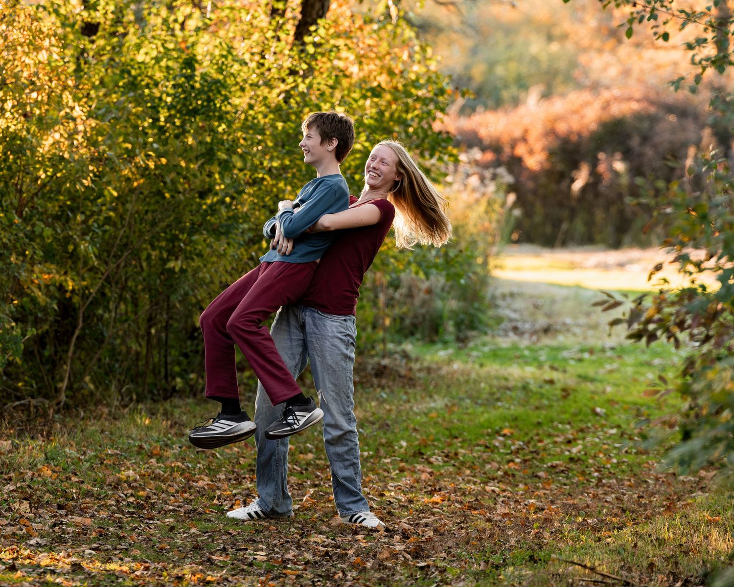 A smiling young woman lifts a laughing boy off the ground in a sunny park, surrounded by golden autumn trees and fallen leaves.