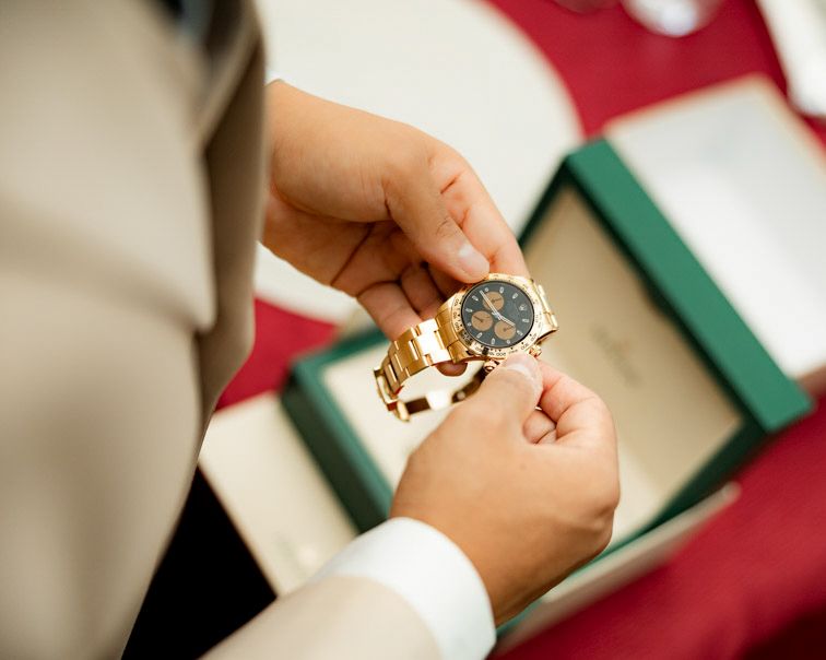 Close-up of hands fastening a gold bracelet watch with a dark dial beside an open green gift box on a red table.