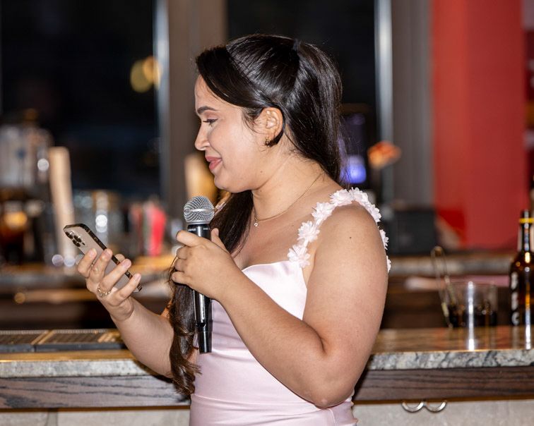 Woman in a blush pink dress with ruffled shoulder detail holds a microphone and phone while giving a speech at an indoor bar venue.