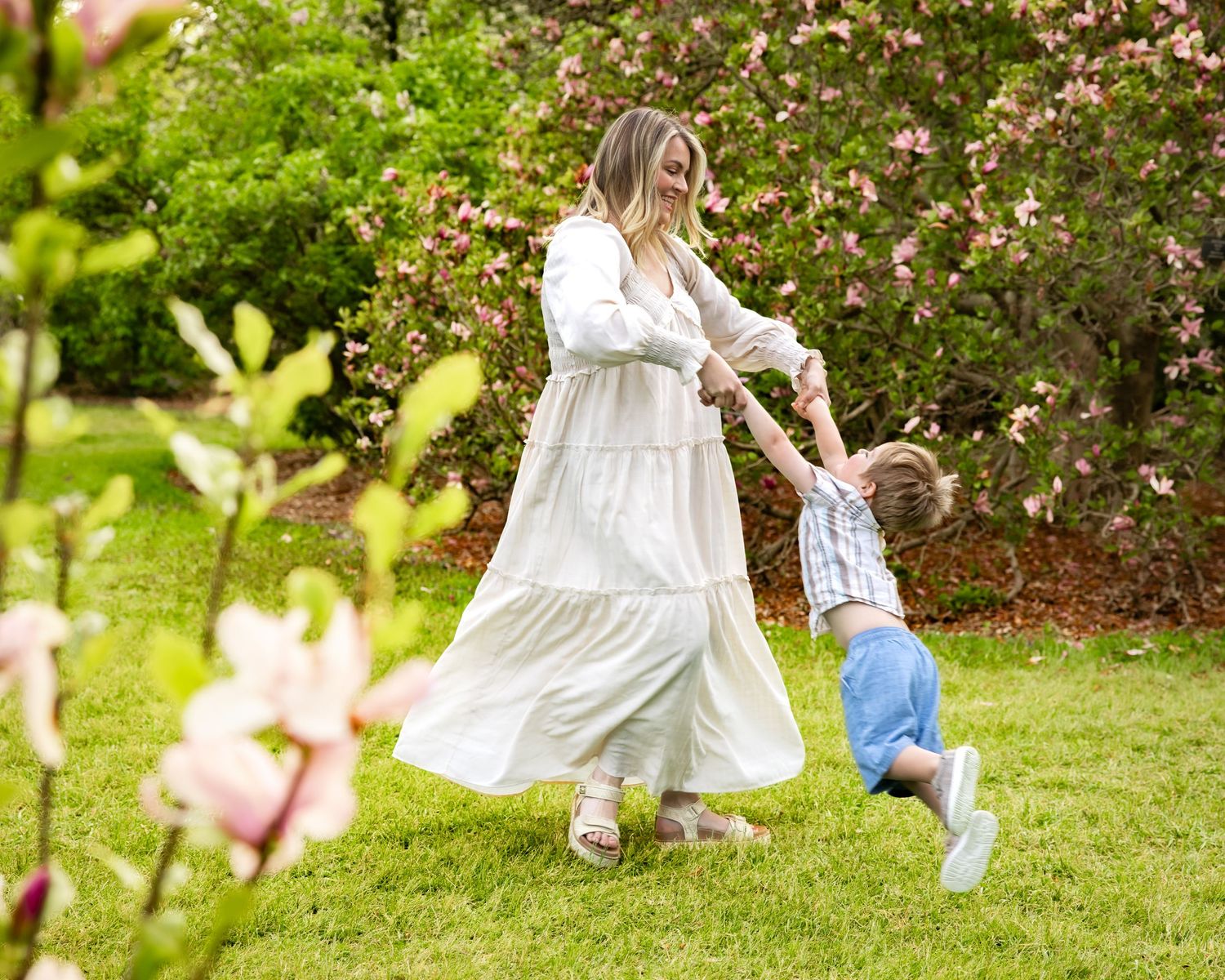 Woman in white dress swinging toddler boy in sunny garden with pink flowering bushes.