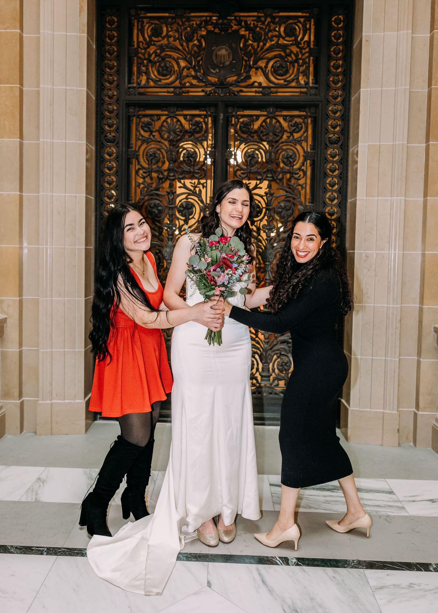 Bride in white dress with colorful bouquet posing with two women before ornate golden doors.