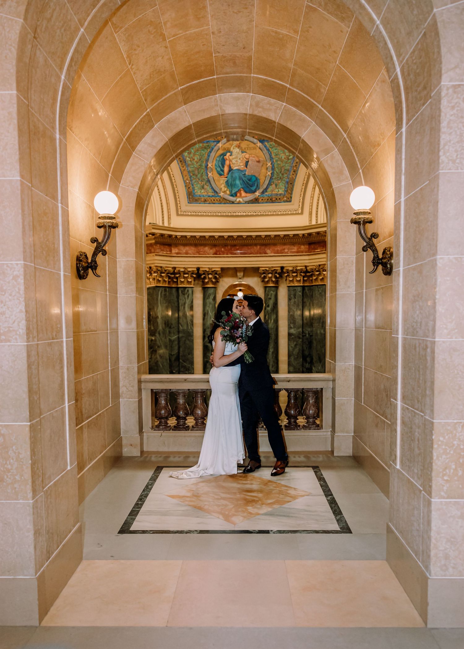 Bride and groom embracing under a beautiful stone arch with colorful mosaic and ornate wall sconces in historic venue.