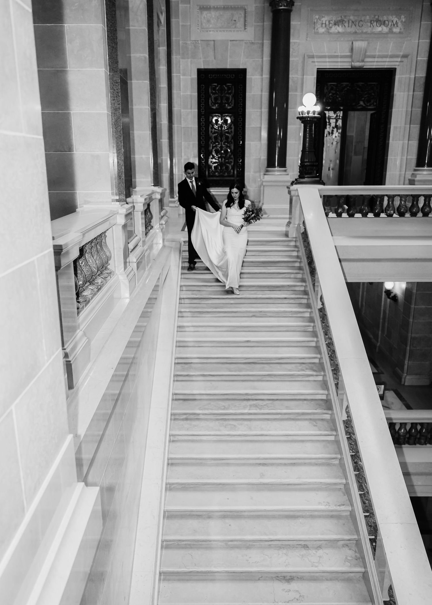 Black and white aerial view of bride and groom descending grand marble staircase together.