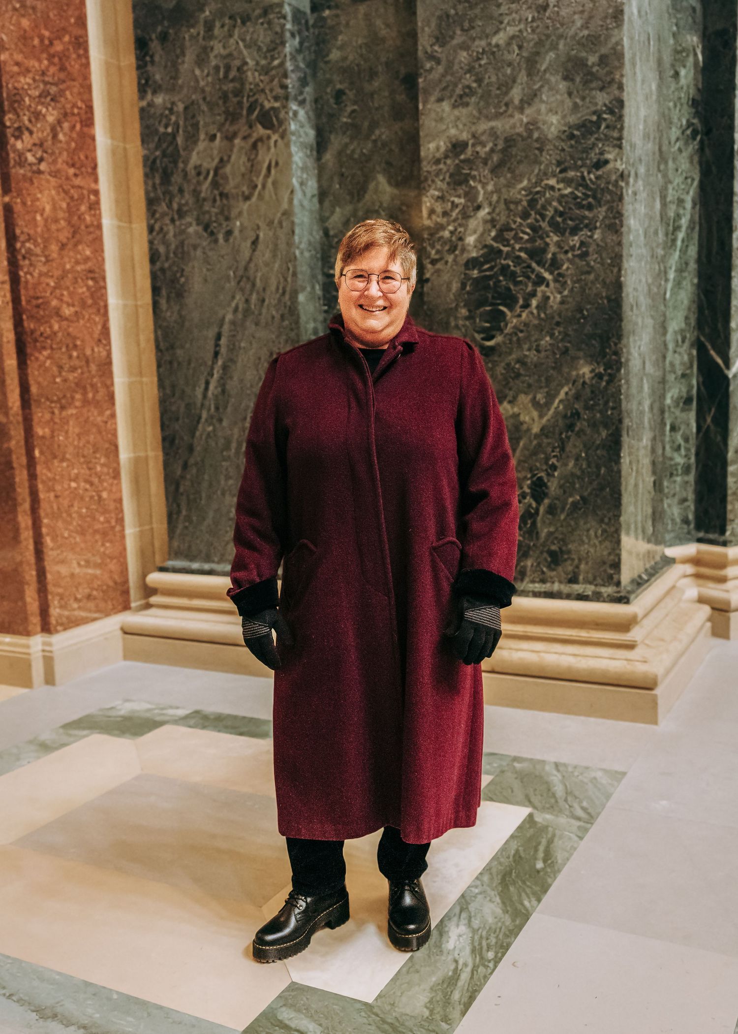 A woman in a burgundy long coat and black gloves smiles between ornate marble columns.