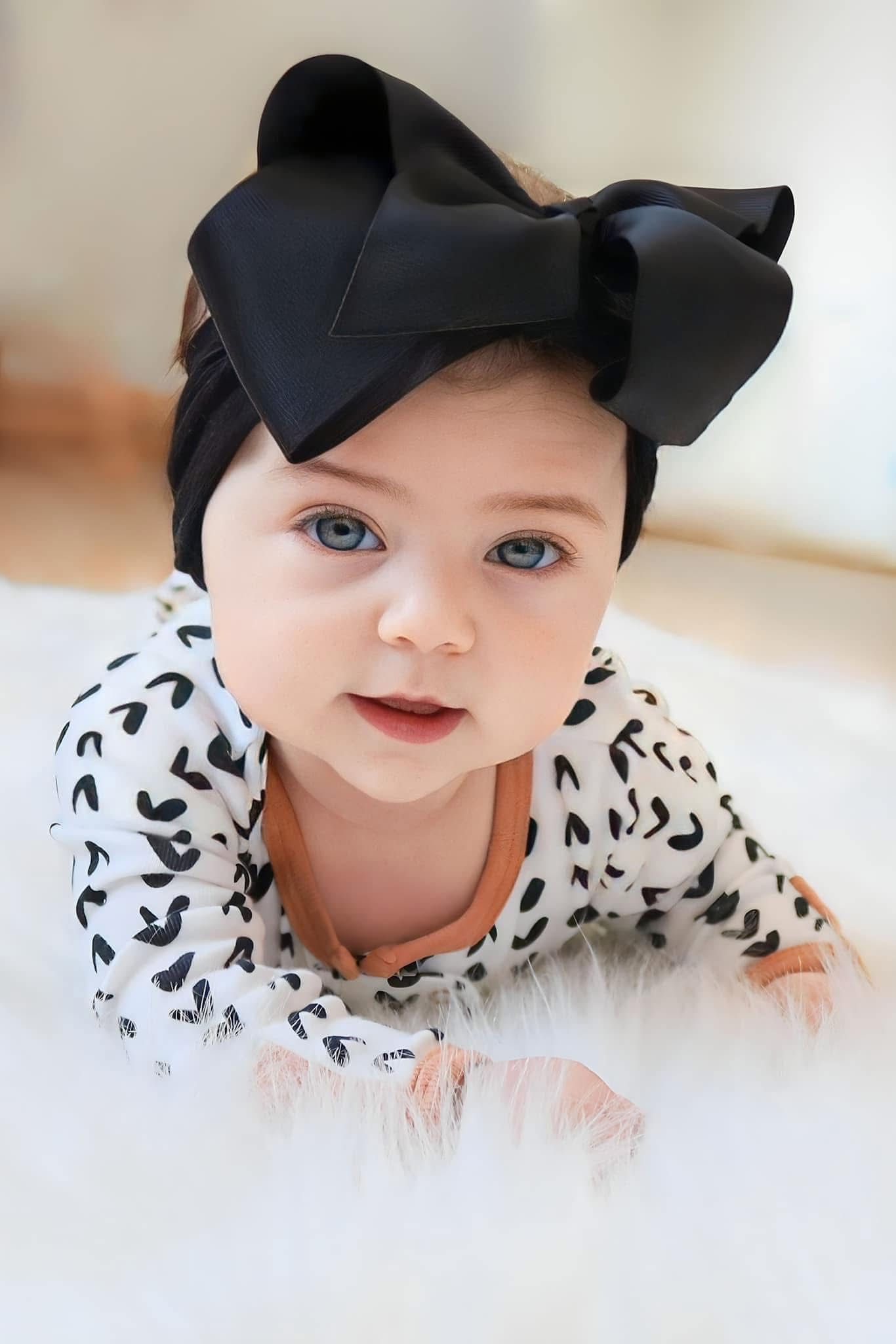 Blue-eyed baby girl in black and white print outfit wears large black bow headband on white rug.