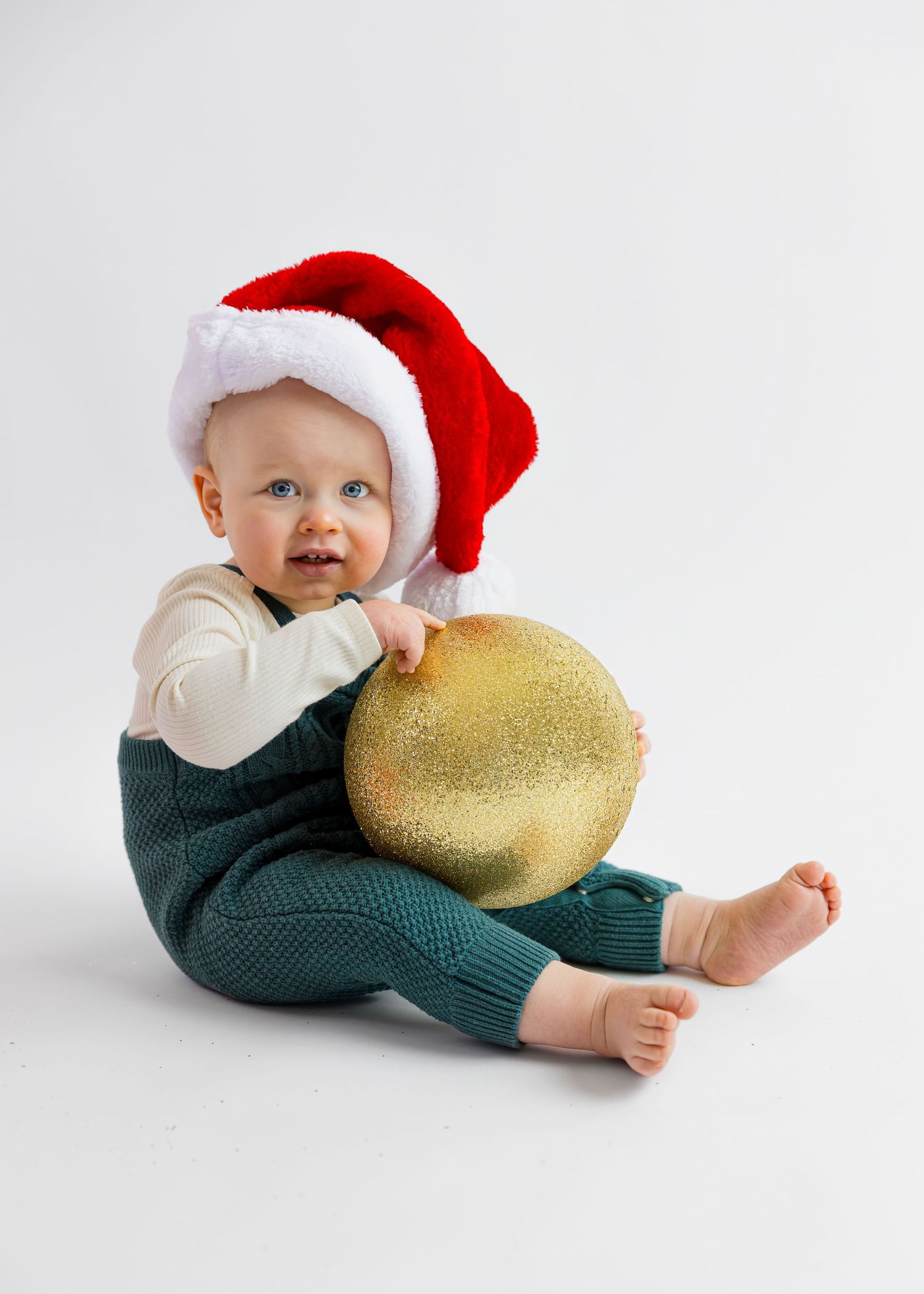 Adorable baby in Santa hat hugs a large gold glitter Christmas ornament while sitting.