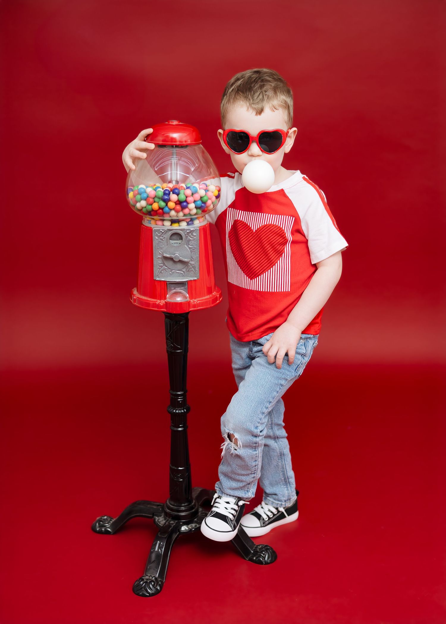 Cool boy in heart sunglasses blowing bubble gum while posing with red gumball machine.