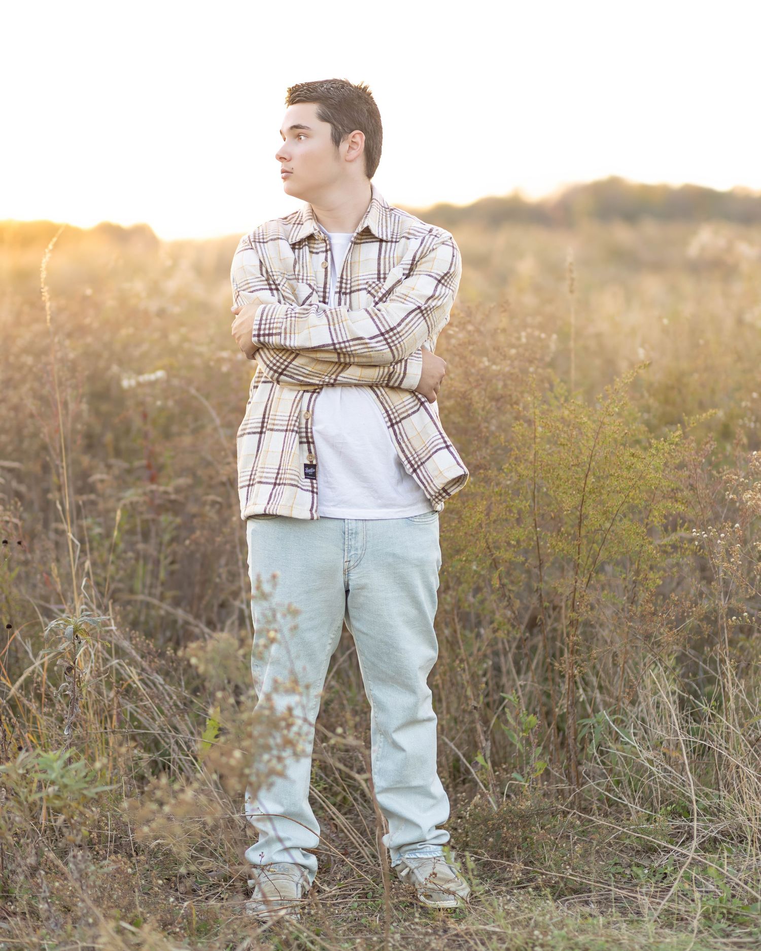 Teen boy in plaid shirt stands with arms crossed in golden autumn field.
