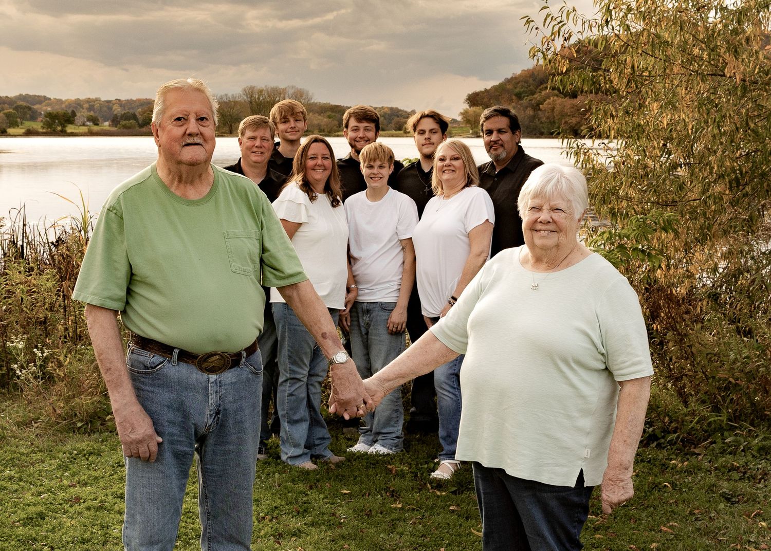 An elderly couple holds hands in front of a multigenerational family group near a scenic lake outdoors.