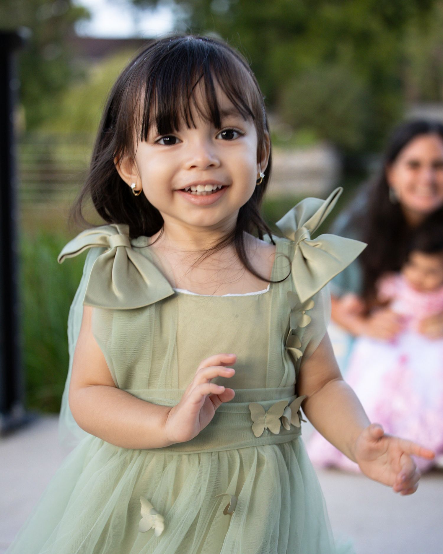 Smiling toddler girl with dark bangs in a sage green tulle dress with bow shoulders and butterfly appliqués, walking outdoors.