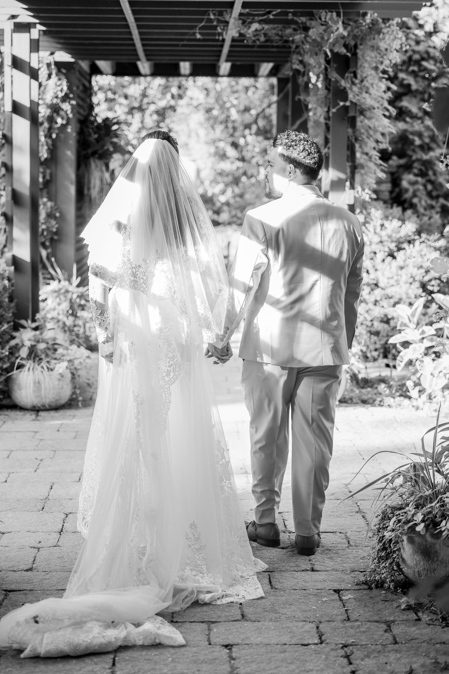 Black and white photo of a bride in a lace gown and veil holding hands with a groom as they walk under a garden pergola.