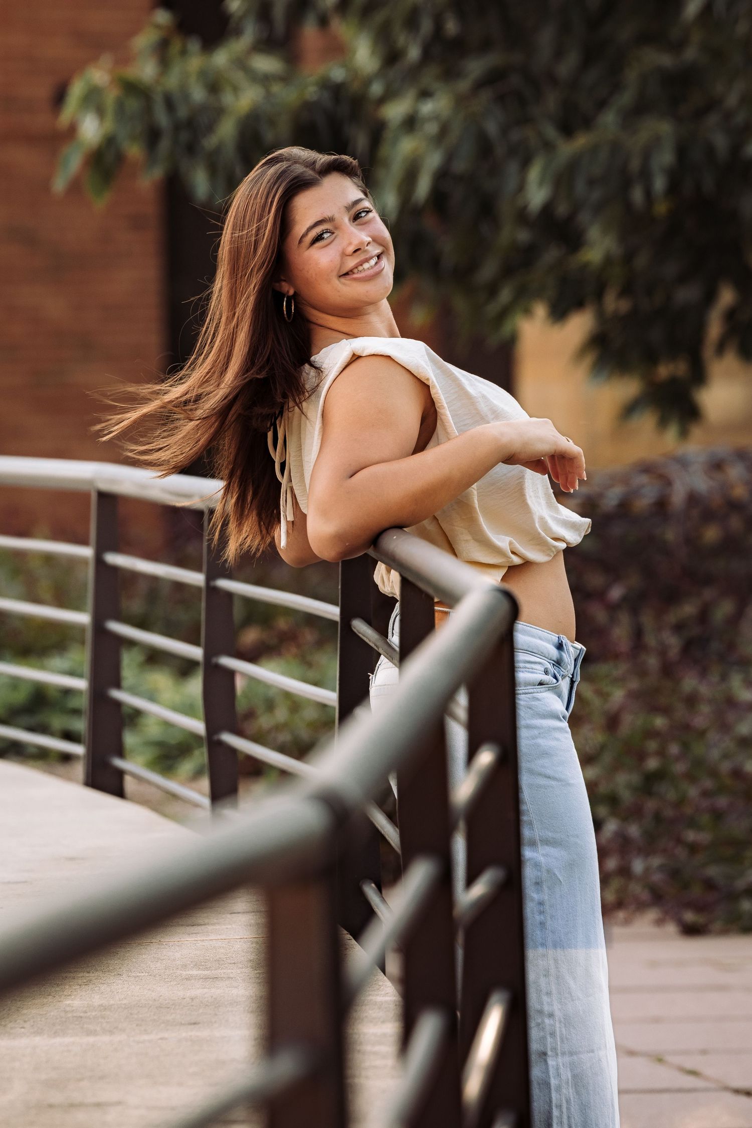 Smiling young woman with long brown hair leaning on metal railing outdoors, wearing beige top and blue jeans.