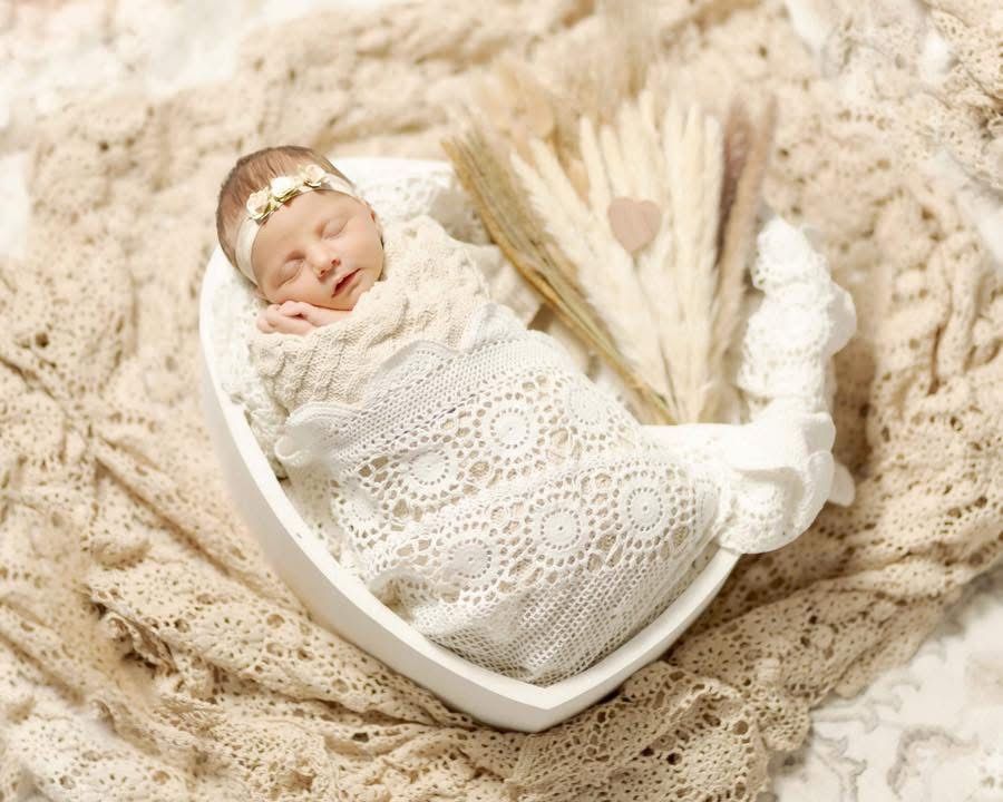 Sleeping newborn baby wrapped in cream lace swaddle in a heart-shaped bowl with dried florals.