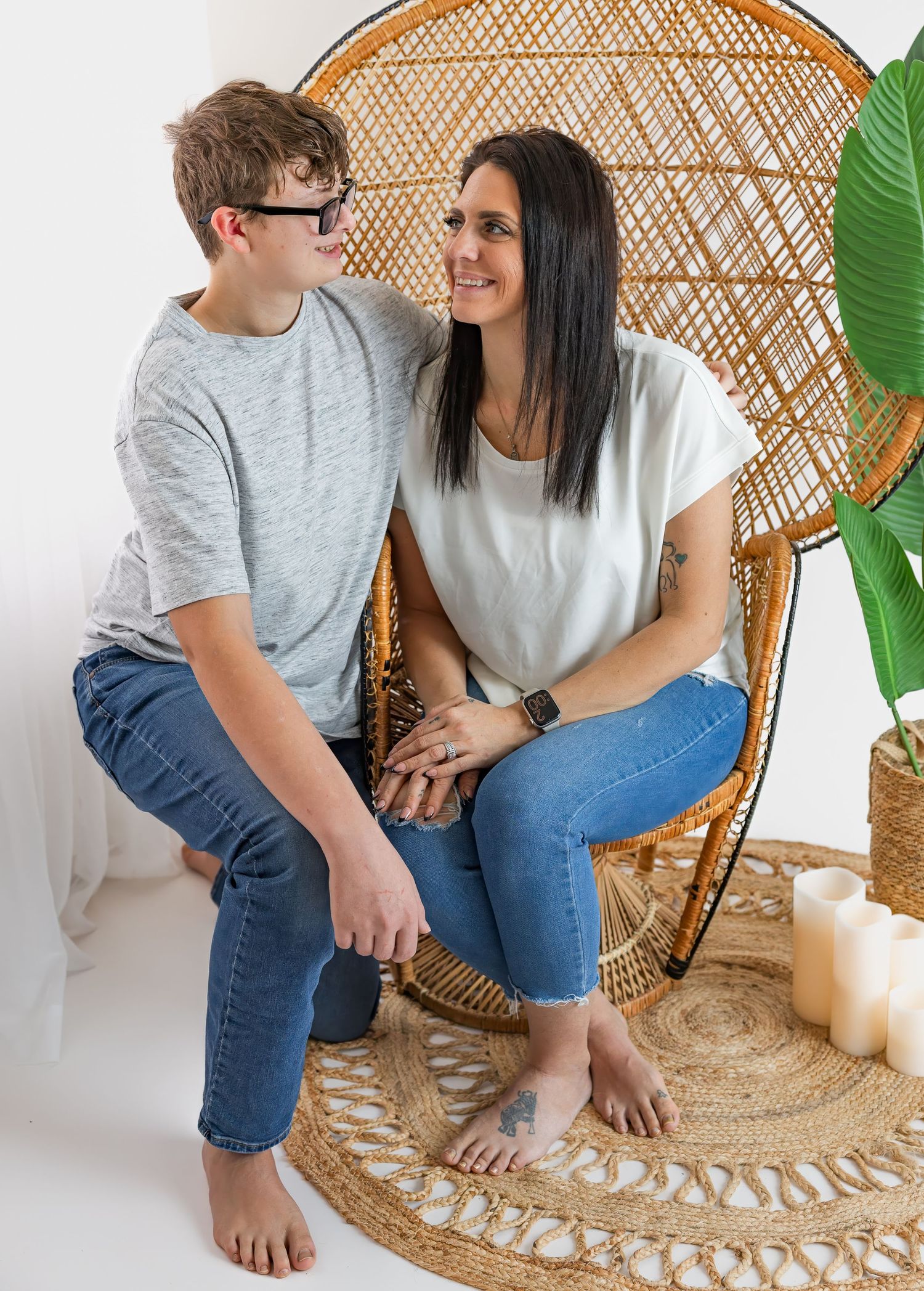 Dark-haired mother and teenage son smiling at each other beside rattan peacock chair indoors.