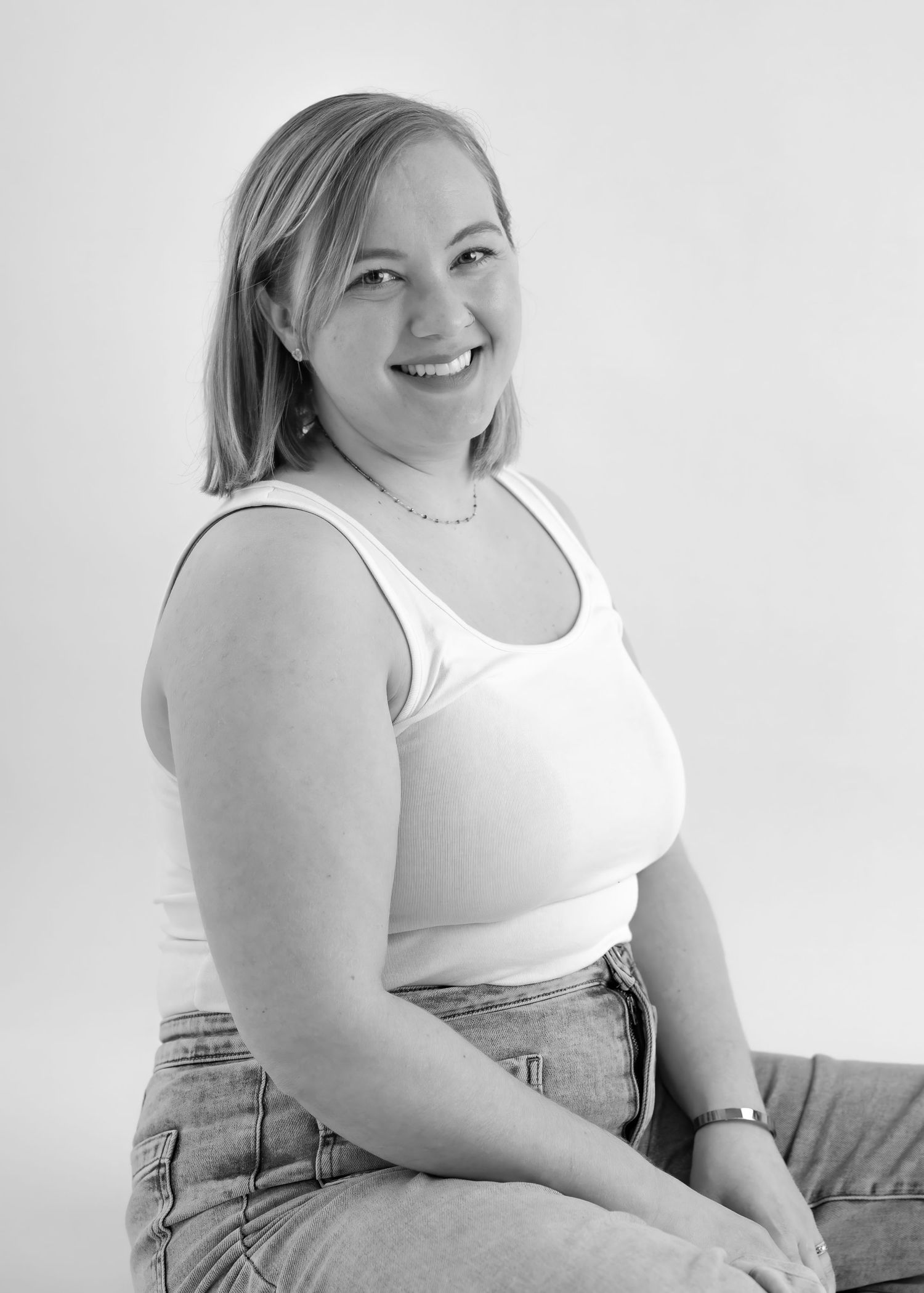 Smiling blonde woman in white tank top and jeans posing in black and white portrait photo.