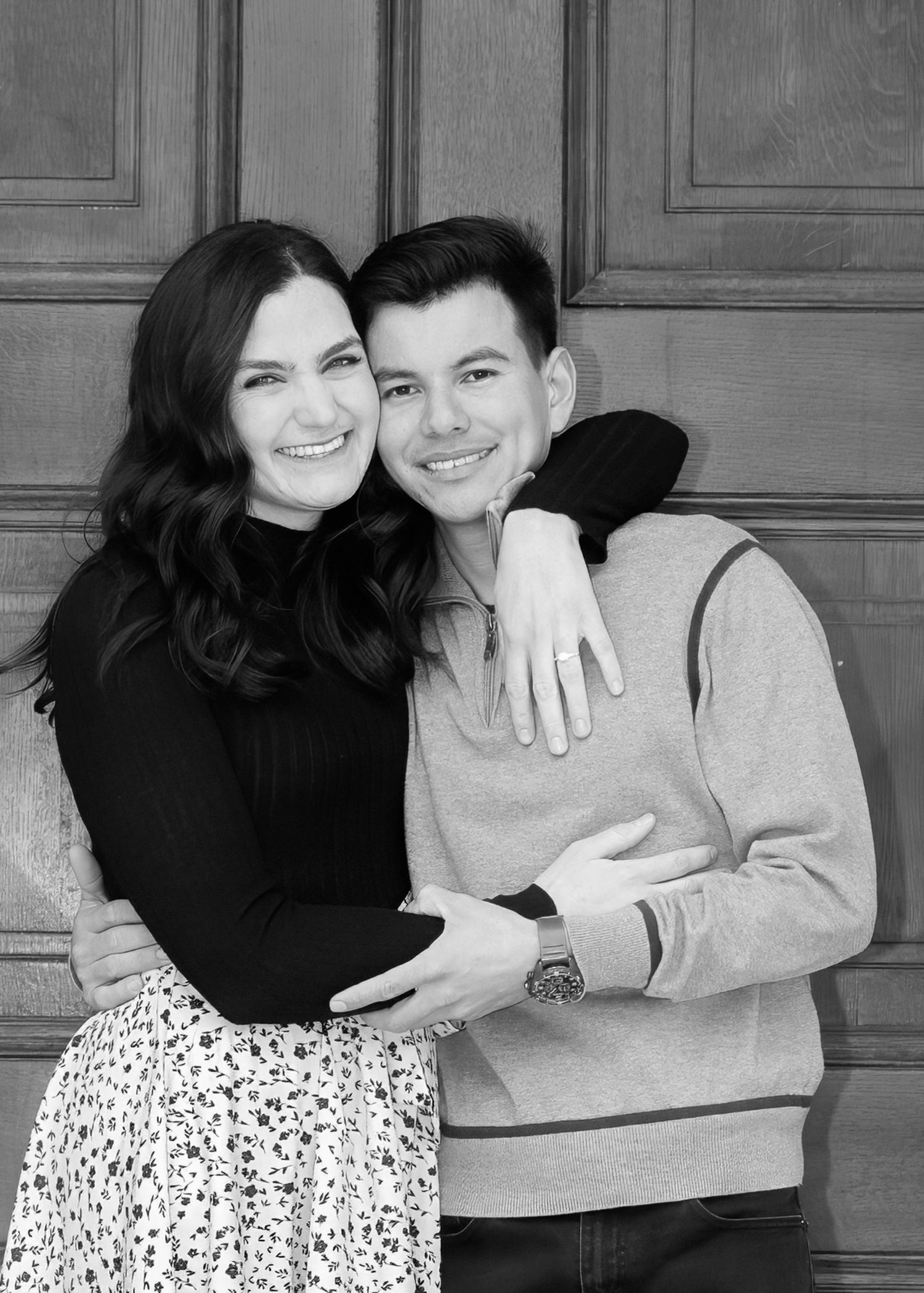 Black and white portrait of smiling couple embracing in front of a wooden door, woman showing engagement ring.