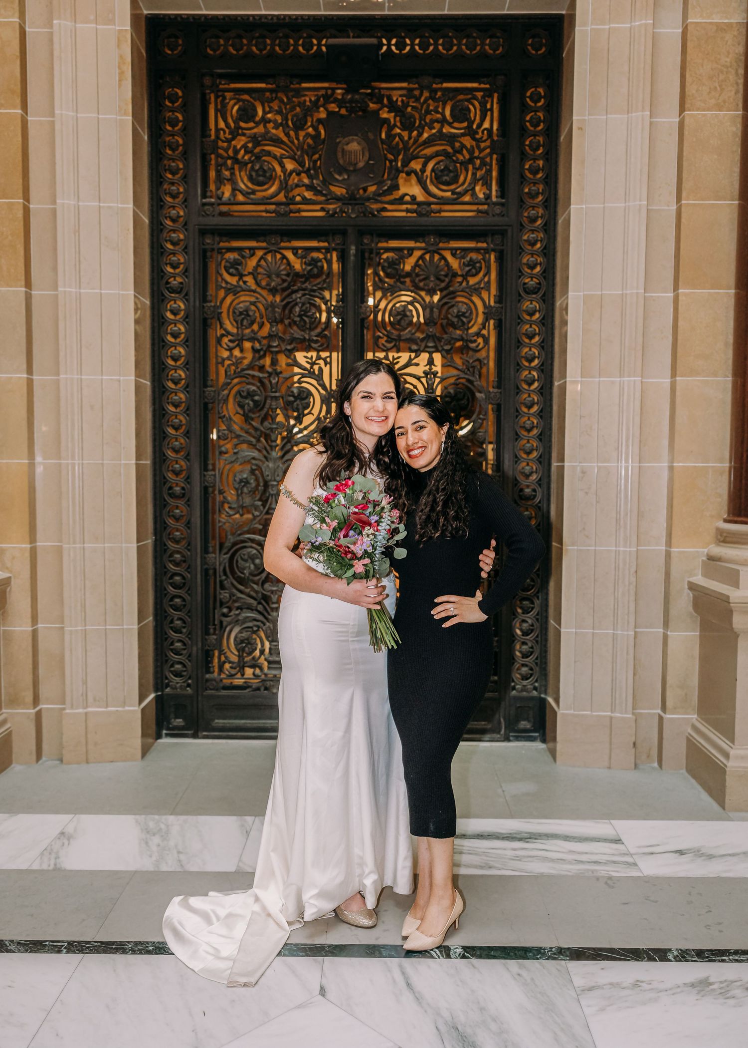 Bride in white gown holding bouquet smiles with guest in black outfit before ornate golden doors.