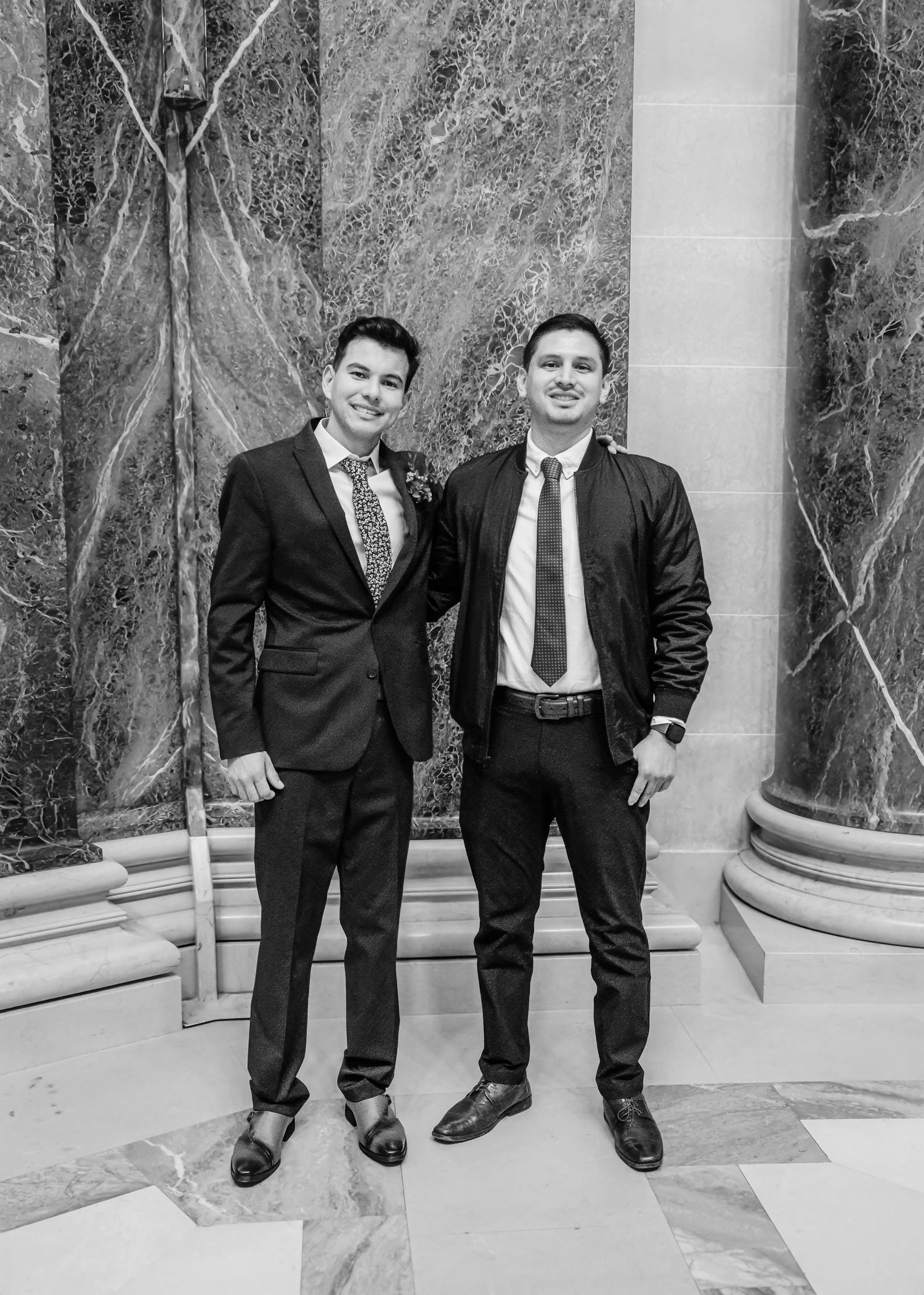 Two well-dressed men in suits and ties posing together in a marble-columned building in black and white.