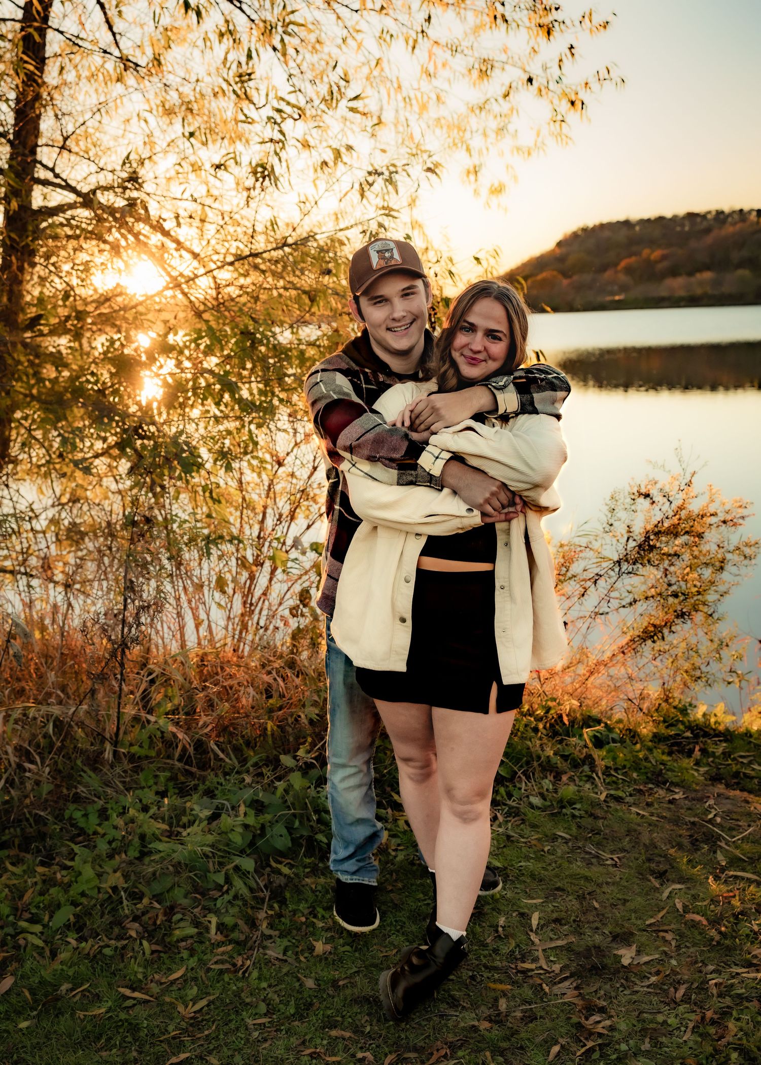 Happy couple hugging and smiling by a lake with golden autumn sunset behind them.