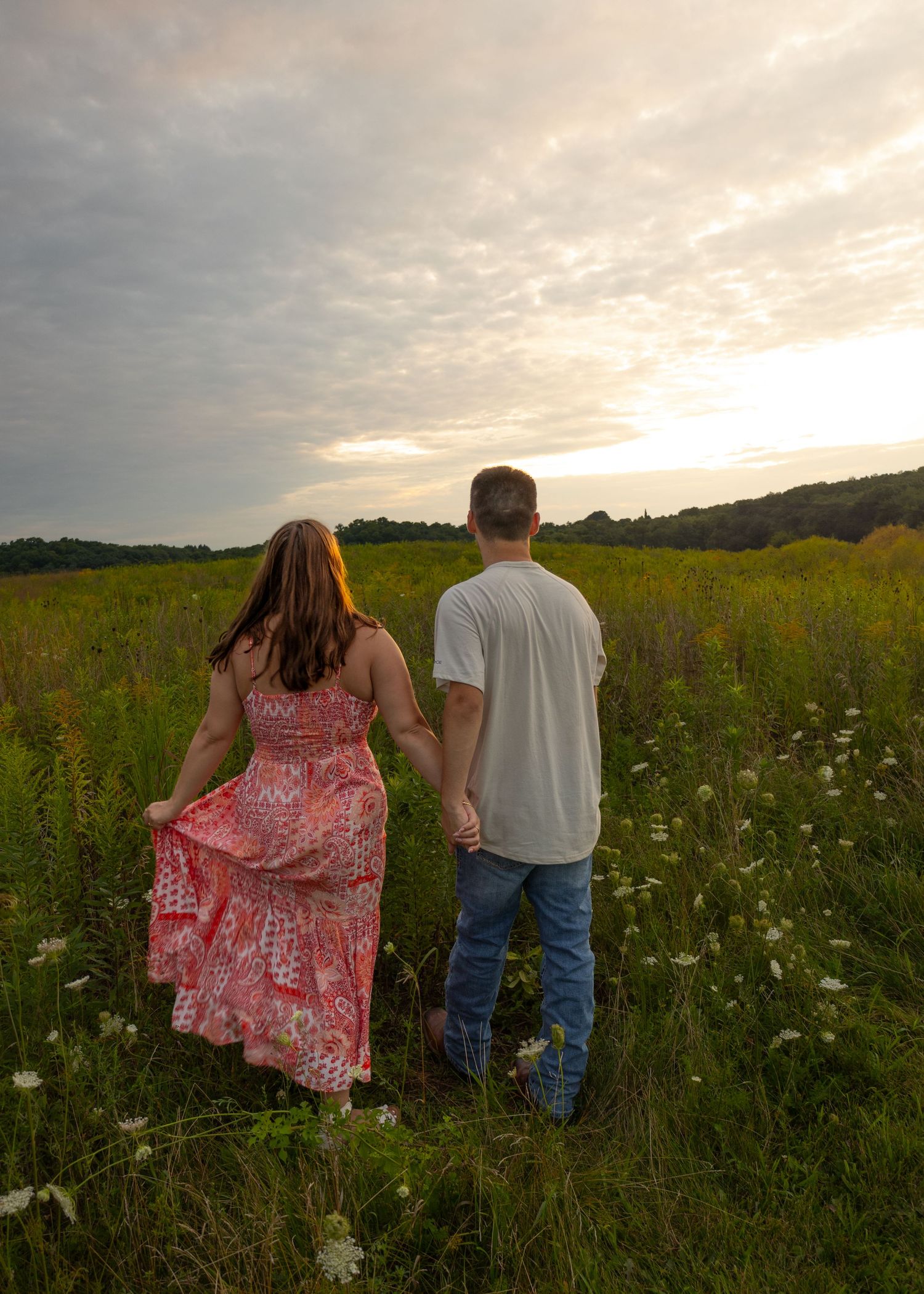 Couple holding hands gazing at a stunning sunset over a lush green wildflower meadow, viewed from behind.