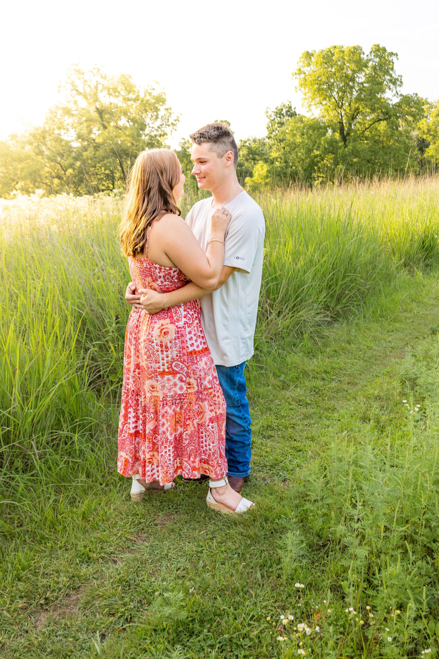 Couple embracing face to face on a wildflower meadow path surrounded by tall green grass at sunset.